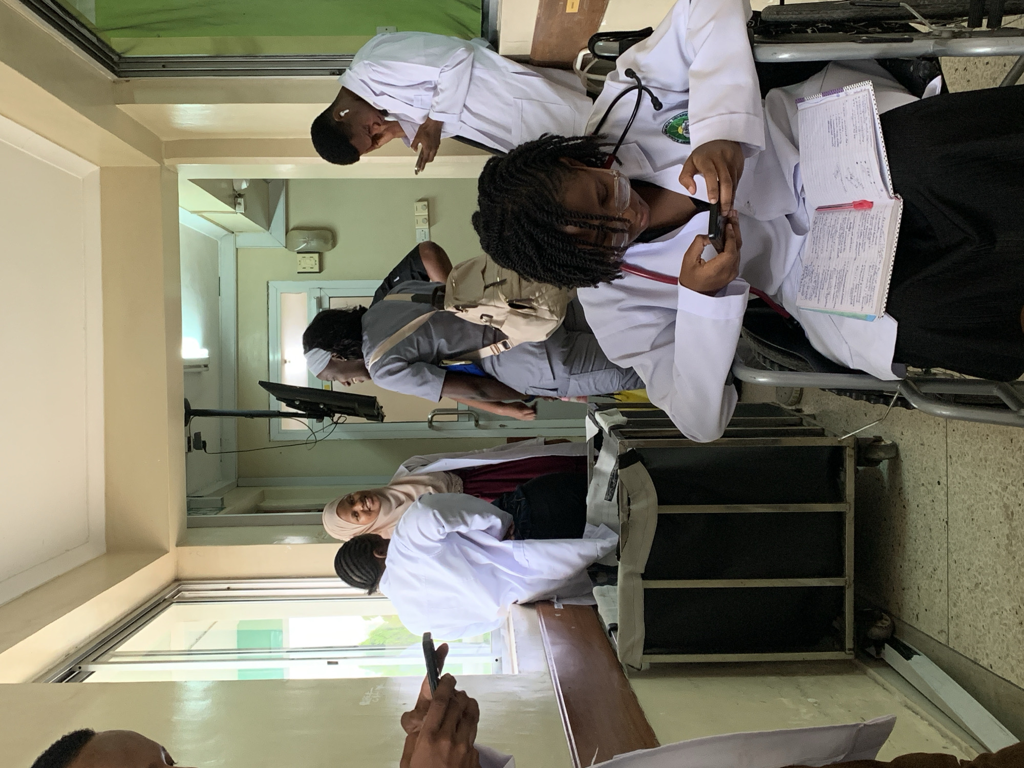In a hallway, presumably inside a hospital or clinic in Dar es-Salaam, Tanzania, several individuals are present. Most appear to be medical professionals or students, identifiable by their white coats and stethoscopes. One person is sitting in a wheelchair, holding a phone in one hand and a notebook with written notes in the other. Another individual is using a phone. Other people in white coats and normal clothes are gathered in the hallway. The environment appears to be a well-lit and busy area.