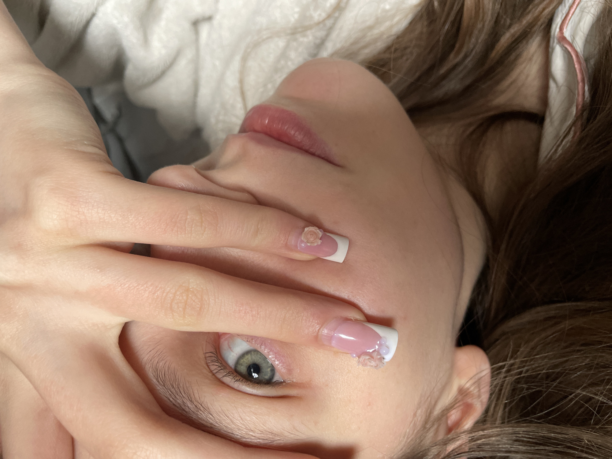 A close-up shot features a person's face and hand. The individual has light skin, green eyes, and brown hair. Their hand is positioned partially over their face, obscuring some features. Two nails are visible, with a light pink base and white tips, decorated with small floral designs. The background is blurred, revealing a light-colored fabric. The location, Фоки, Russia, is not discernible from the visual information provided.