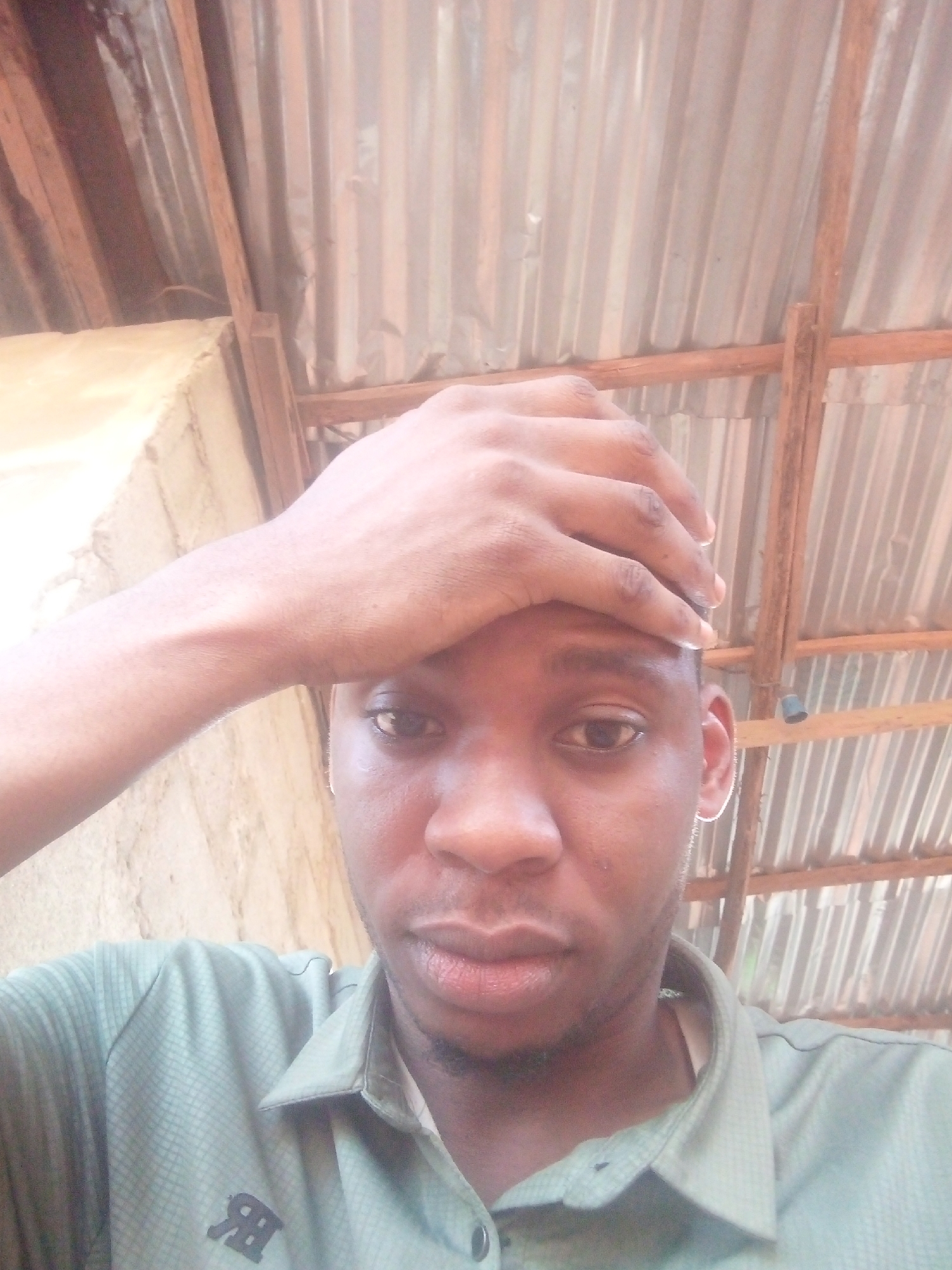 A medium-shot selfie of a dark-skinned person with short, dark hair. They are wearing a light green button-up shirt with a logo resembling the letters "R" and "B" on the left side of the shirt. The person's right hand is placed on their forehead. The background consists of corrugated metal roofing supported by wooden beams. A wall with a rough, light-colored surface is visible on the left side of the frame. The location is in the city of Unknown, Unknown.