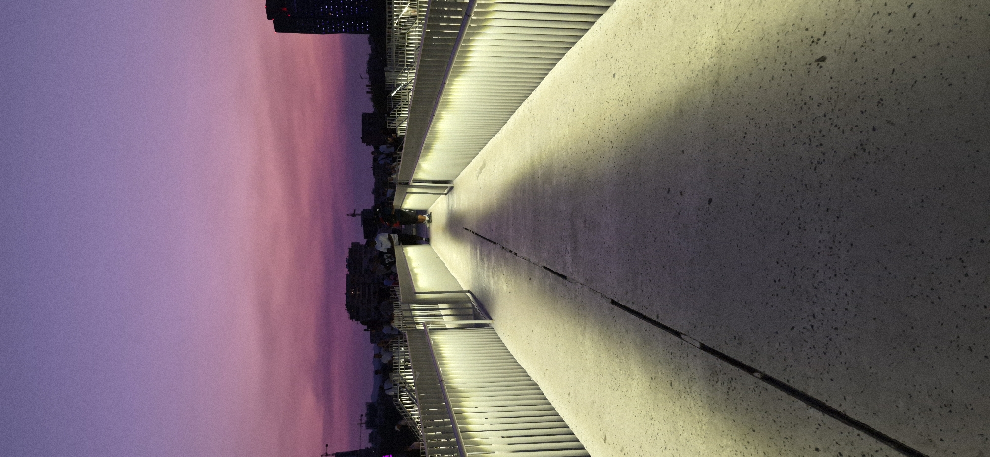 The image depicts a modern pedestrian bridge at dusk in Tirana, Albania. The sky is a gradient of deep purple at the top, transitioning to pink and lighter purple hues towards the horizon. The bridge itself is made of concrete and is illuminated by vertical, soft white lights along its railings, casting long shadows onto the walkway.
In the background, the city skyline is visible with several buildings, including a tall, dark skyscraper on the right side. Construction cranes are also present in the distance, suggesting ongoing development. Several people are scattered across the bridge, some walking and others standing, appearing to be enjoying the view or an event. Two individuals are clearly visible in the foreground, one standing and looking towards the distance, and another walking away from the camera. The bridge has a wide walkway with a visible seam running down the center. The overall atmosphere is calm and modern, capturing a moment of urban life during twilight.