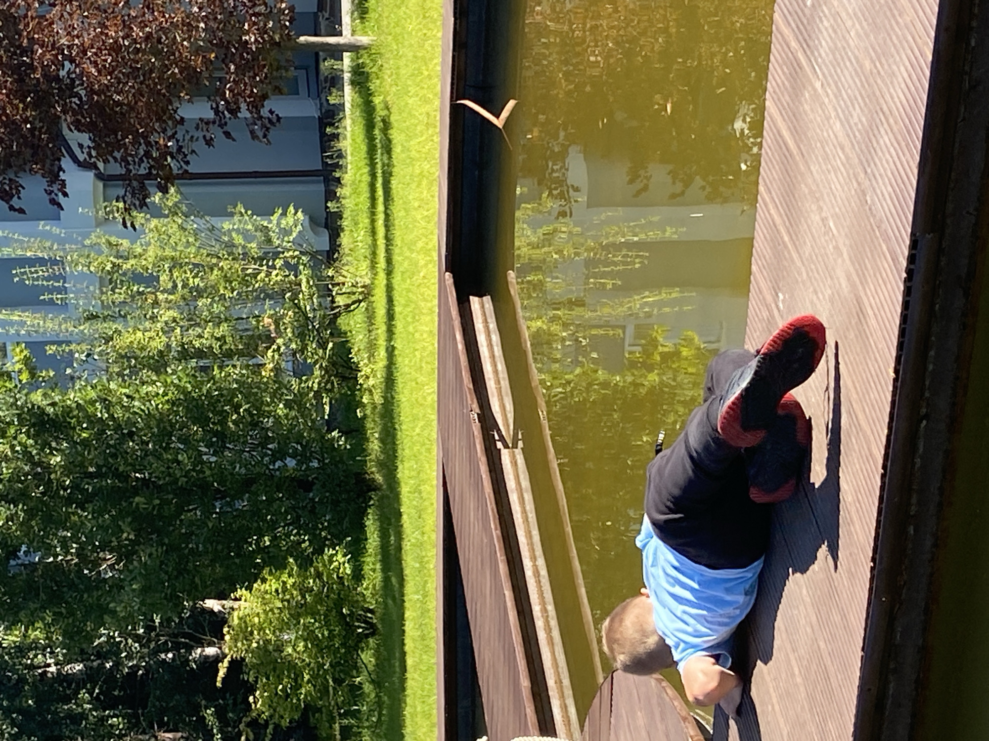 A young boy wearing a blue shirt and black pants with red-soled shoes lies on a wooden deck next to a pond. The boy is facing away from the camera, looking into the murky green water. The deck appears to be part of an outdoor area with lush green grass and trees in the background, suggesting a park or residential setting. A building with a light-colored facade is visible behind the trees. The lighting indicates it is daytime, with sunlight casting shadows on the deck. The location is identified as Chulis, Russia.