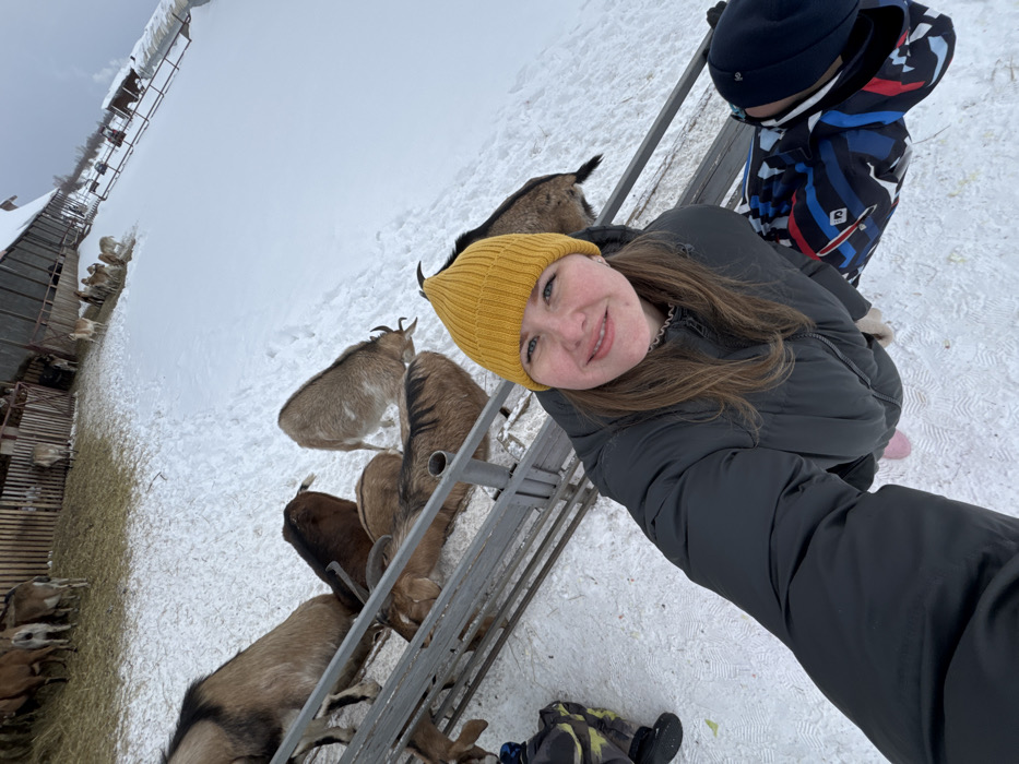 An overhead-angle image depicts an outdoor winter scene at a goat farm in Zaosinovo, Russia. The foreground features a smiling adult female wearing a yellow knit beanie, a dark grey padded winter jacket, and light pink footwear, holding a camera to take a selfie. Her light brown hair is visible beneath the beanie. To her right and partially obscured is a child wearing a dark blue and black patterned winter jacket and a dark blue beanie. Both individuals are positioned on a snow-covered ground adjacent to a metal barrier or fence.
Beyond the barrier, several domestic goats of varying brown, tan, and grey coloration are present on the snow. Some goats are directly adjacent to the barrier, while others are further into the enclosure. In the middle ground, more goats are visible within a fenced-off area with hay on the ground. The background features a rustic wooden barn structure and additional fenced areas, all surrounded by snow. The sky is overcast, indicating a winter day.