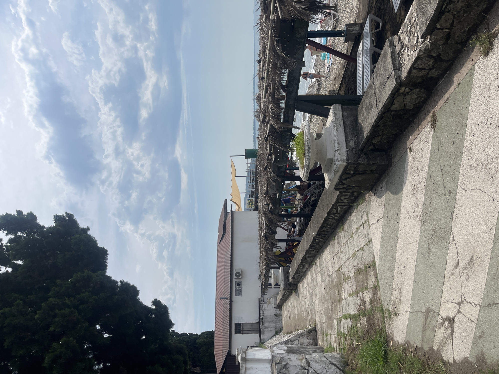 The image depicts a view of a beach in Sokhumi, Abkhazia, on a cloudy day. In the foreground, a paved walkway with visible cracks and patches of grass runs alongside a stone wall. The wall supports a covered area with thatched roofs, under which lounge chairs and some people are visible. Beyond this area is a pebble beach with more lounge chairs and a few people. The sea stretches out to the horizon under a sky filled with scattered clouds. To the left of the walkway is a white building with a red-tiled roof, featuring an air conditioning unit and a barred window. A large, dark green tree dominates the upper left portion of the image.