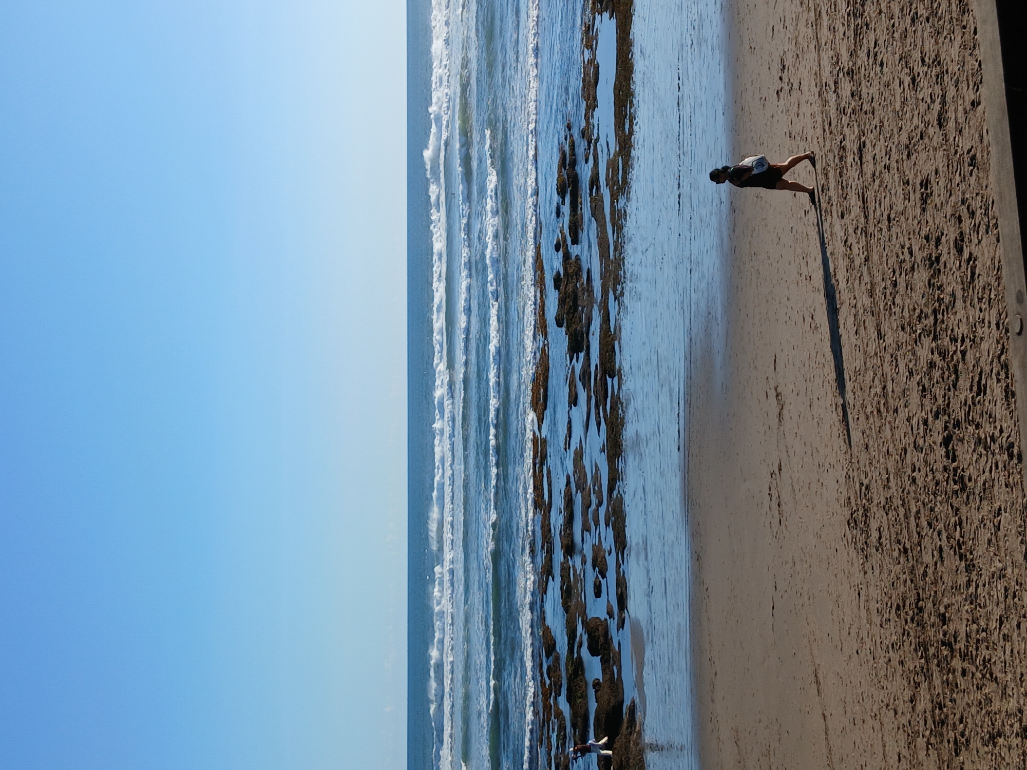 An eye-level, medium shot captures a serene beach scene under a clear blue sky in North Kuta, Indonesia. The foreground is dominated by a sandy beach with textured patterns, suggesting recent foot traffic or wind action. In the mid-ground, a solitary woman walks across the sand, her silhouette cast long and dark by the sun. She is wearing dark clothing and carrying a light-colored bag over her shoulder. The middle ground transitions to the ocean, where gentle waves with white crests roll towards the shore. A rocky intertidal zone, partially submerged in the water, lies between the beach and the open sea. In the far distance, the horizon separates the deep blue ocean from the vast expanse of the bright blue sky.
