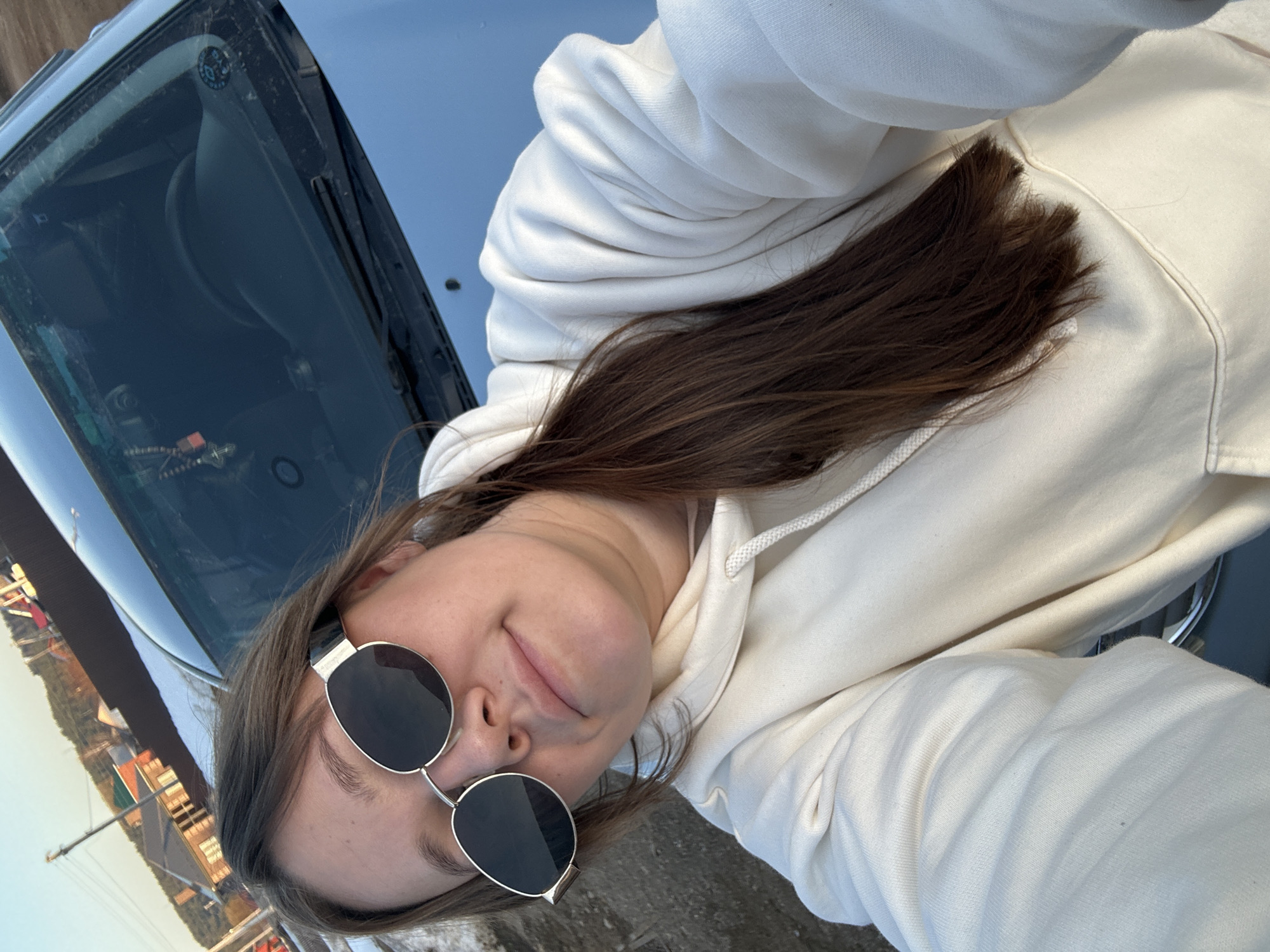 A woman with long brown hair is wearing round sunglasses and a white hoodie. She is leaning against a dark-colored car, possibly taking a selfie. The edge of a body of water and some buildings are visible in the background, suggesting an urban or coastal location. It is difficult to confirm the specific location of Ольховка, Russia, without further information.