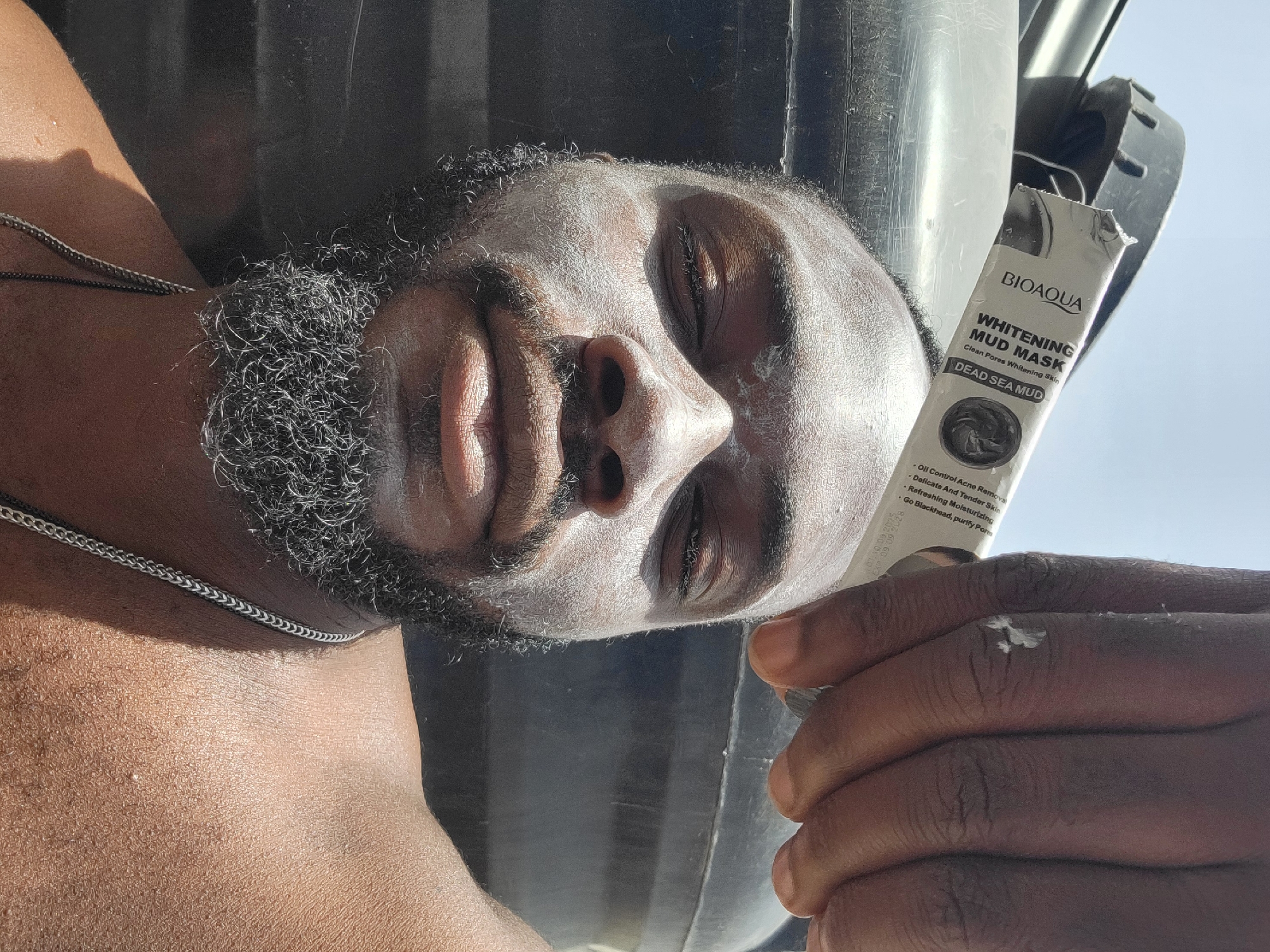 A dark-skinned man with a beard and a coating of white mud mask on his face lies supine, holding a white and black tube of Bioaqua Whitening Mud Mask in his right hand. A drop of the mud mask is visible on the back of his hand. He wears a thin silver chain around his neck. The background is unclear, but elements suggest an outdoor setting, possibly in Jos, Nigeria, based on the geographical setting requested in the prompt.