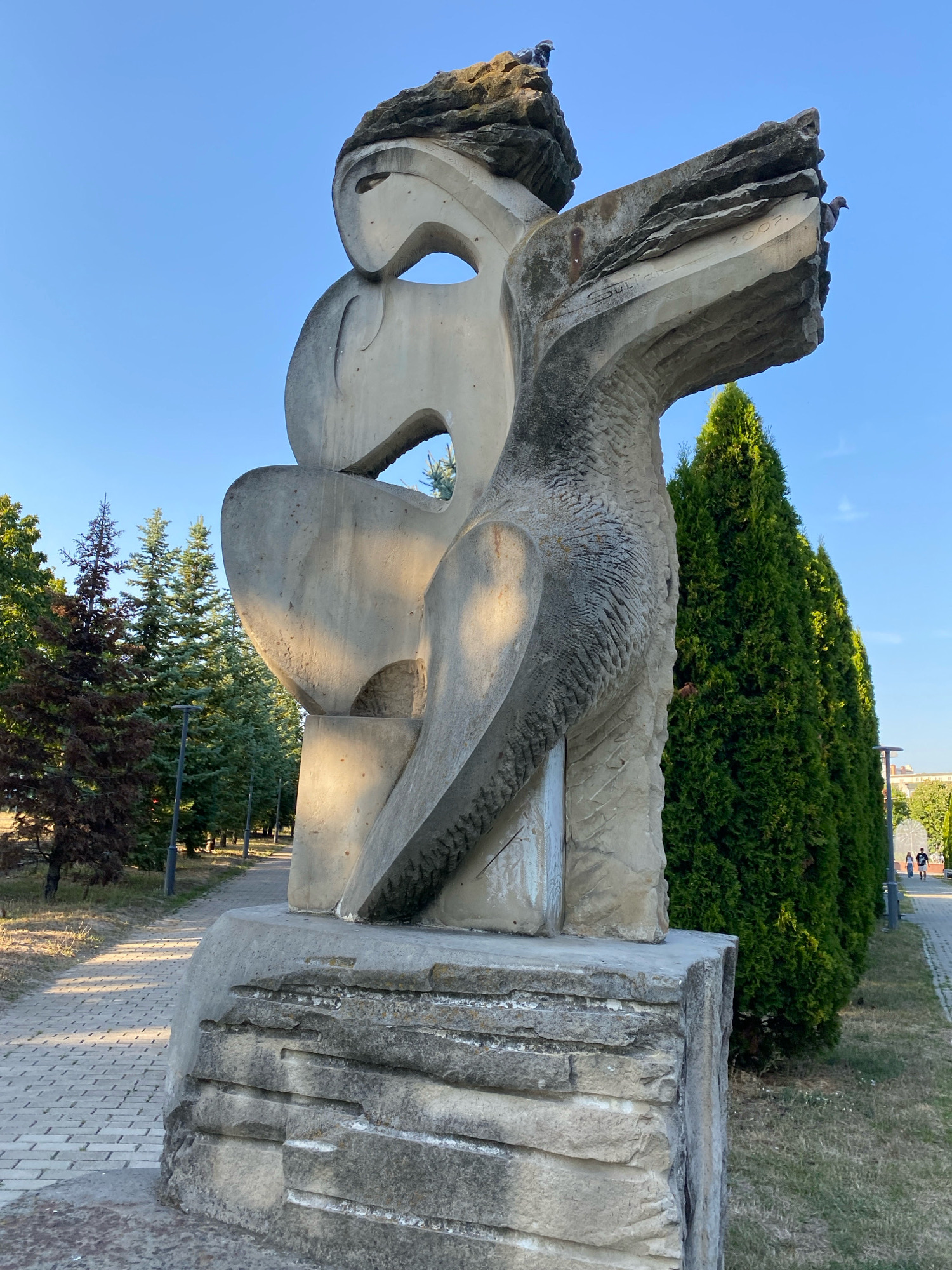 The image shows a large, abstract stone sculpture in a park-like setting. The sculpture is light gray and appears to be made of several large, rough-hewn stone blocks assembled together. It has an organic, almost bird-like form with sharp angles and curves. There is an inscription on the sculpture that reads "2007". Two pigeons are perched on the upper part of the sculpture.
The sculpture is situated on a paved walkway with a stone border. To the left of the sculpture, there are several conifer trees, some of which appear to be dry or brown. Further in the background, there is a row of tall, green cypress trees. The ground is a mix of grass and dry earth. In the distance, to the right of the cypress trees, a few people can be seen walking on a path. The sky is clear and blue.
This sculpture is located in the Alley of Classics in Chișinău, Moldova.