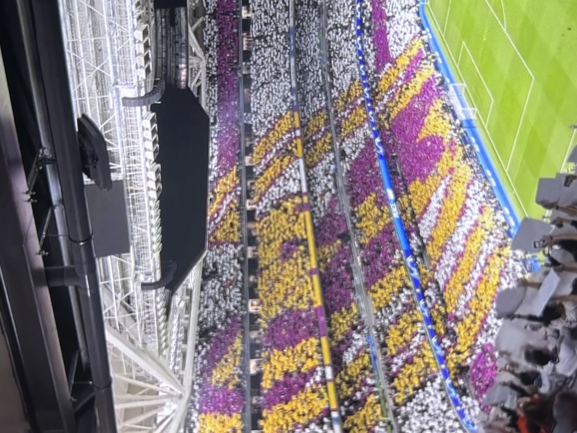 An aerial, rotated view captures a large, modern sports stadium filled with a crowd during a night event. The stands are packed with people, many holding up colored cards or lights to create a vibrant tifo display. This organized spectacle features alternating horizontal and diagonal stripes of purple, yellow, and white, covering a significant portion of the stadium seating.
Visible stadium infrastructure includes large support beams, metal framework, and what appear to be speaker arrays or screens suspended above the stands. A section of a bright green football (soccer) pitch with white markings and a goal is discernible in the upper right.
The artificial lighting illuminating the stadium and field indicates the event is taking place at night. The coordinated visual display suggests an enthusiastic and united atmosphere, likely in support of a team or performer at a sporting event. Given the location context of Bulgaria, the purple and yellow colors are notably associated with CSKA Sofia, a major Bulgarian football club. No clear text is visible on banners or screens.