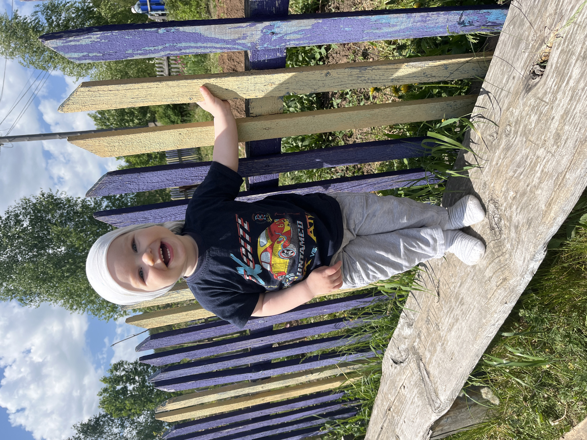 The image shows a toddler in Сосново, Russia, lying on a weathered wooden plank leaning against a painted wooden fence. The fence planks are alternating colors of purple and yellow. The toddler is wearing a white cap, a dark t-shirt with a car graphic, and grey pants with white socks. The toddler is smiling and holding onto the fence. Green grass and foliage are visible around the plank and fence. The sky is visible through the trees and fence, with scattered clouds. A building is visible in the background to the left of the fence.