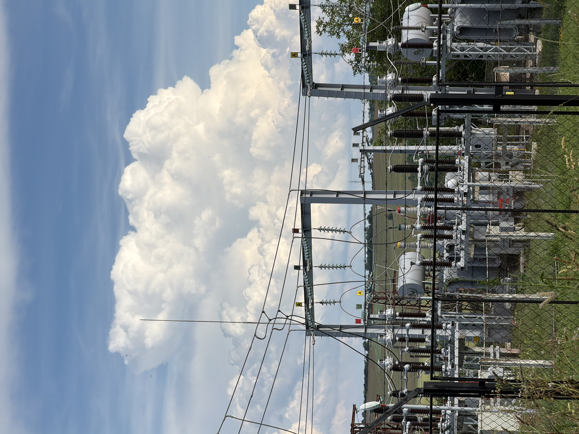 The image depicts an electrical substation with large, white cumulus clouds dominating the sky in the background. The substation features numerous electrical transformers, insulators, and thick cables supported by metal structures. A chain-link fence runs across the foreground, with dry grass and weeds growing around it. In the distance, a rural landscape with rolling hills and scattered trees can be seen under the partly cloudy sky. The specific location of this substation is not provided in the image itself.