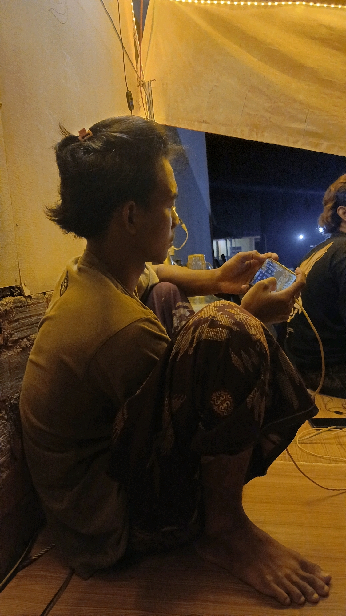 An individual, identified as a male by his physical characteristics, is depicted in a seated position, facing to the right, and engrossed in playing a mobile game on his smartphone. The individual is wearing a light brown t-shirt and a patterned sarong that covers his lower body. His hair is styled in a short bun at the back of his head, secured with a pink hair tie. He has a yellow wire extending from his right ear, possibly connected to earbuds.
The setting appears to be outdoors, possibly in a relaxed or casual environment. Behind the individual, a wall with a textured surface is visible. Above, a large, light-colored fabric canopy is stretched, adorned with a string of small, warm-toned lights, suggesting it is evening or nighttime. The background also reveals glimpses of buildings and the night sky. The ground is made of wooden planks, and electrical wires are visible, some yellow and one red. Another individual, partially obscured and wearing a black t-shirt, is visible to the right. The overall ambiance is one of casual recreation in an outdoor setting at night, illuminated by artificial lighting. The location is specified as Adi Luhur, Indonesia.