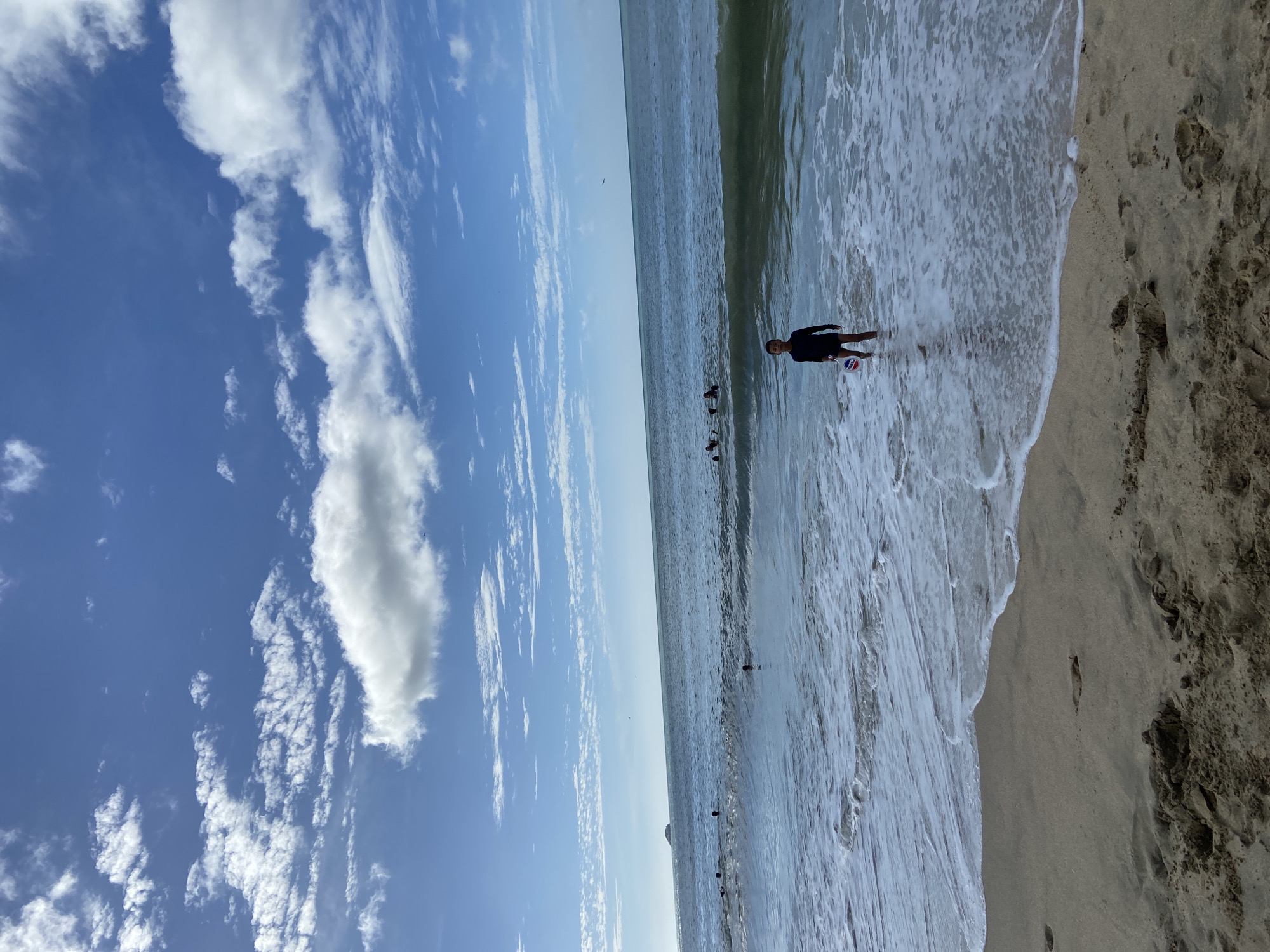 A medium-shot, horizontal, outdoor photograph depicts a beach scene in Pedro González, Venezuela. The beach runs from the foreground to the horizon line. The left side of the image is dominated by a sky of blue with scattered cumulus clouds. The right side of the image shows the shoreline, where white waves meet the light-colored sand. A person is standing in the shallow water near the center of the frame, holding a ball. There are other people farther out in the water.