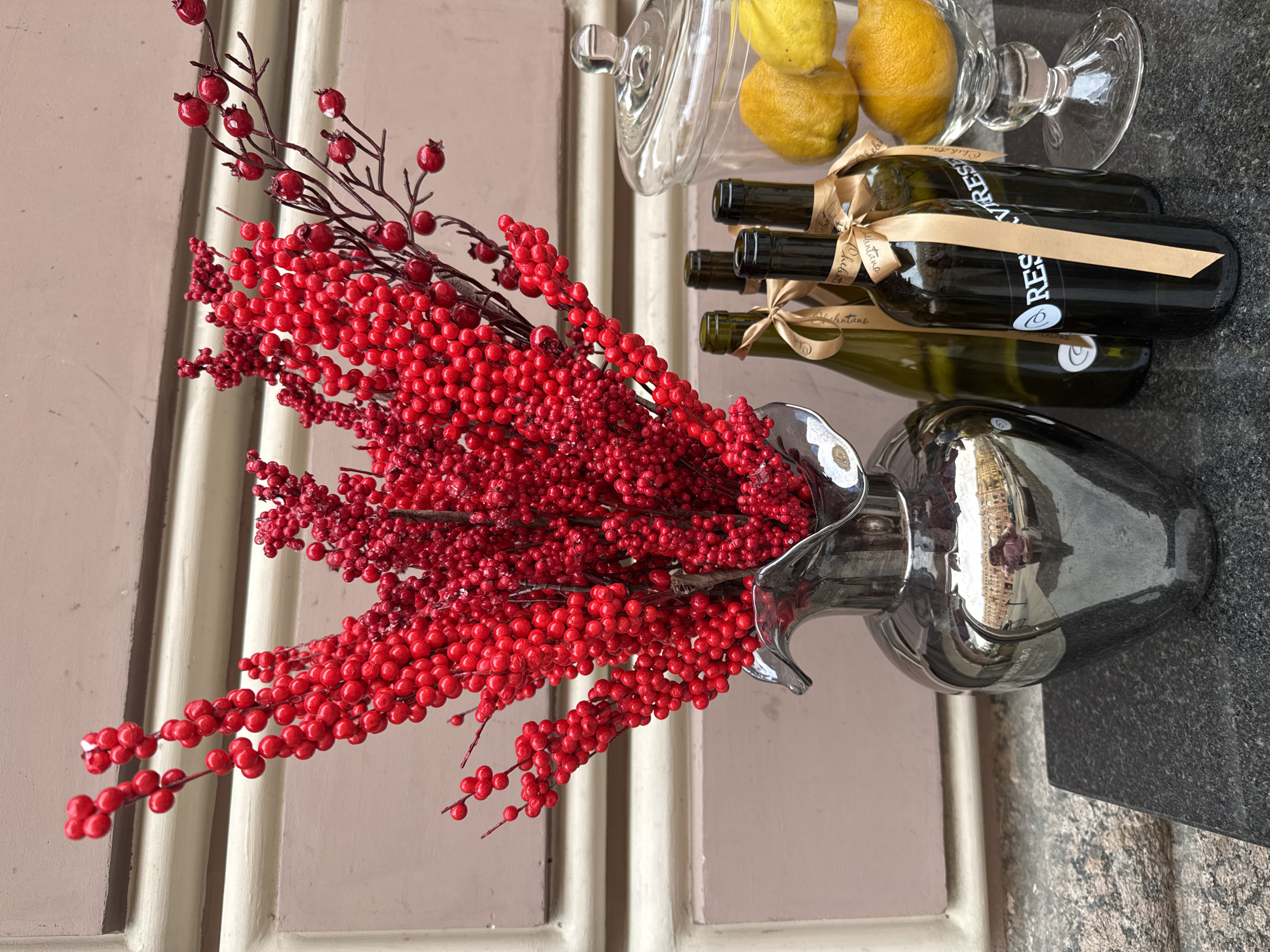 A view of Saint Petersburg.
The photograph captures a silver vase filled with bright red berry branches. Nearby, there are several dark wine bottles, each adorned with a light brown ribbon. A glass container filled with lemons rests next to the bottles. The arrangement sits atop a stone surface adjacent to a wall with vertical panels painted in a neutral color. In the reflection of the silver vase, a building can be observed, suggesting the location is Saint Petersburg, Russia.