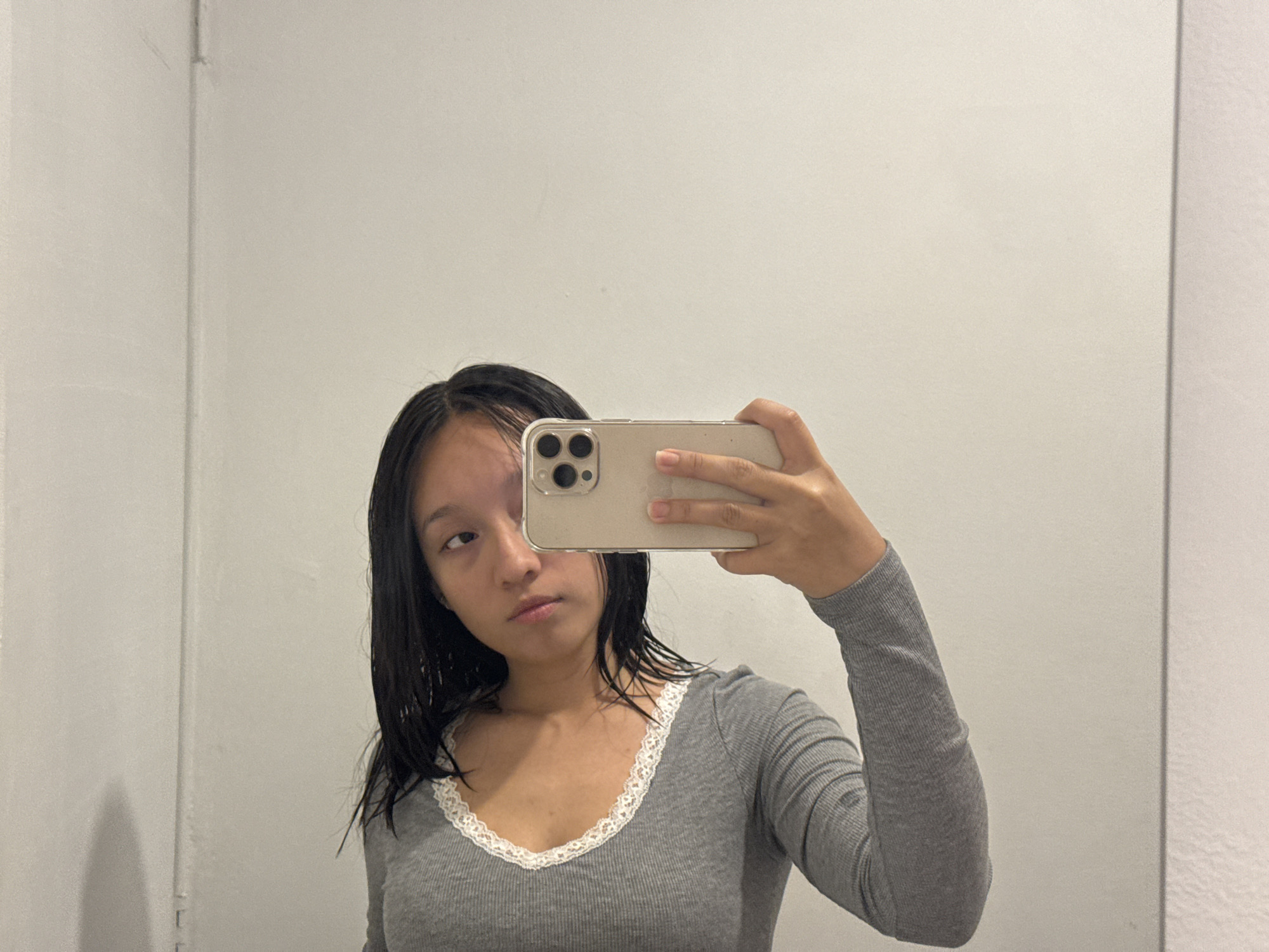 A young woman is taking a selfie with her phone. She is wearing a gray long-sleeved shirt with white lace trim around the neckline. Her hair is dark and appears to be wet. The background is a plain white wall with subtle vertical lines that may indicate a paneled or painted surface. The environment appears to be indoors, possibly in a bathroom or a room with a mirror. The location in the city of Puebla, Mexico, is not discernible from this image.