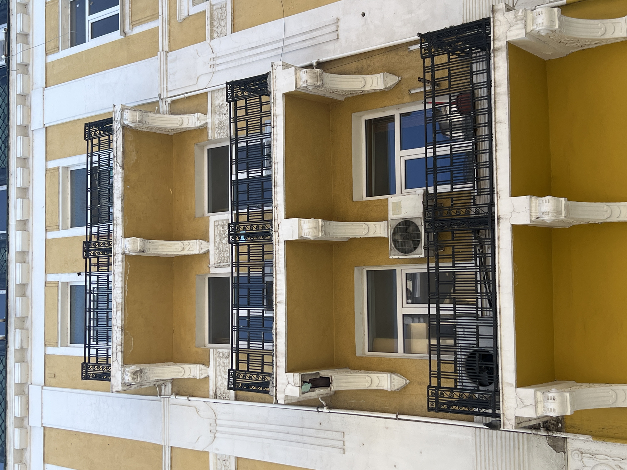 An eye-level, slightly tilted-up shot shows a yellow multi-story building with white accents and ornate protruding ledges. Black metal fire escapes are installed outside several windows. Windows are visible, some with air conditioning units placed inside or outside. The building is located in an urban environment, and the location is Unknown, Unknown.