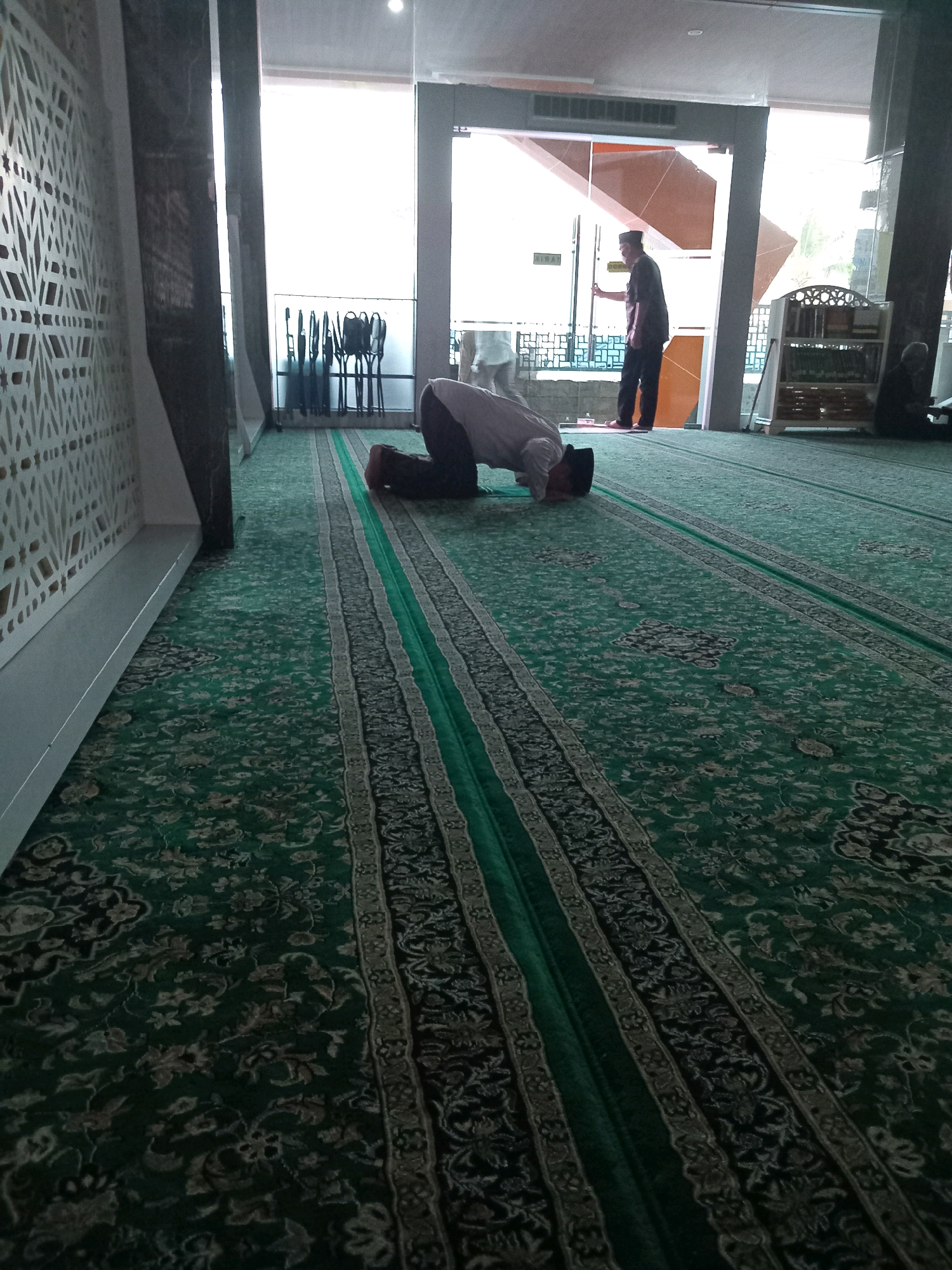 An indoor shot taken from a low angle shows a man in a mosque in Randuagung, Indonesia, performing the Sujud position during prayer. He is wearing a white shirt and a black skullcap, with his head touching the green carpet. The carpet has a repeating floral pattern in shades of green, beige, and black, with distinct rows marking prayer spaces. To the right of the praying man, an upright prayer rug with a darker pattern is visible. In the background, a man in a dark shirt and black pants, wearing a white skullcap, is standing near a glass door and a wooden shelf filled with books. Another person is seated to the far right, also in prayer. The environment appears to be a modern mosque with large windows, letting in natural light.
