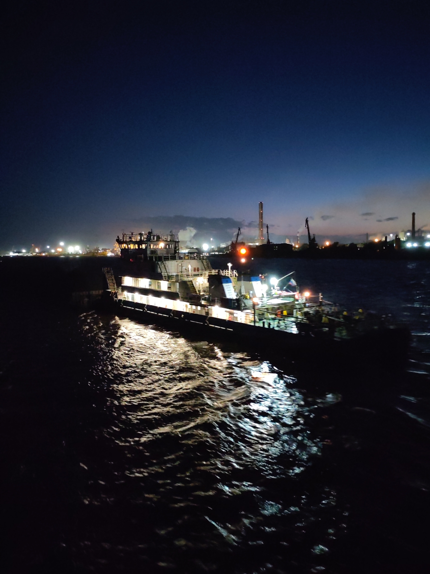 A dimly lit, nocturnal scene shows a barge or tugboat navigating a waterway. The vessel is illuminated with multiple lights, revealing its structure with multiple decks and a cabin or control area on the top level. Reflections of the lights dance on the water's surface. In the background, the silhouetted cityscape features industrial structures like chimneys and cranes, and their lighting suggests this location may be in Cherepovets, Russia. The sky is a dark blue, hinting at twilight or night. There are no clearly visible people in the foreground, but people are seen on the barge.