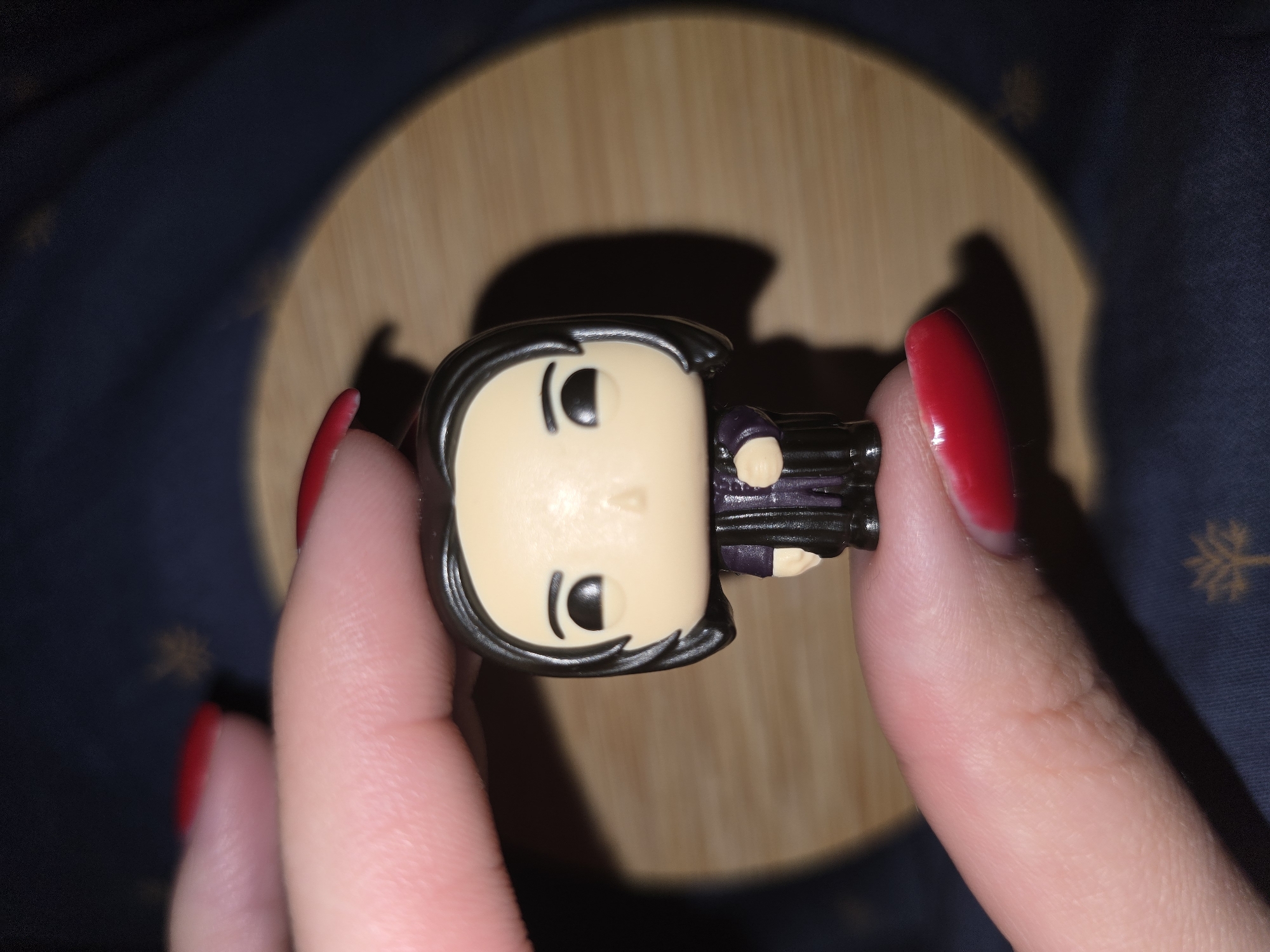 A close-up shot shows a person holding a Funko Pop figurine of a character with dark hair, large eyes, and a purple outfit. The figurine is held horizontally by two fingers, one on each side of its torso. The background is blurred, with a wooden surface and a dark fabric with a subtle snowflake pattern. The lighting creates a shadow of the figurine on the wooden surface. The person's hands and fingernails are visible, with red nail polish on the fingernails. The scene appears to be indoors, possibly in a home environment, given the dark fabric with a decorative pattern. The focus is on the figurine and the hands holding it.