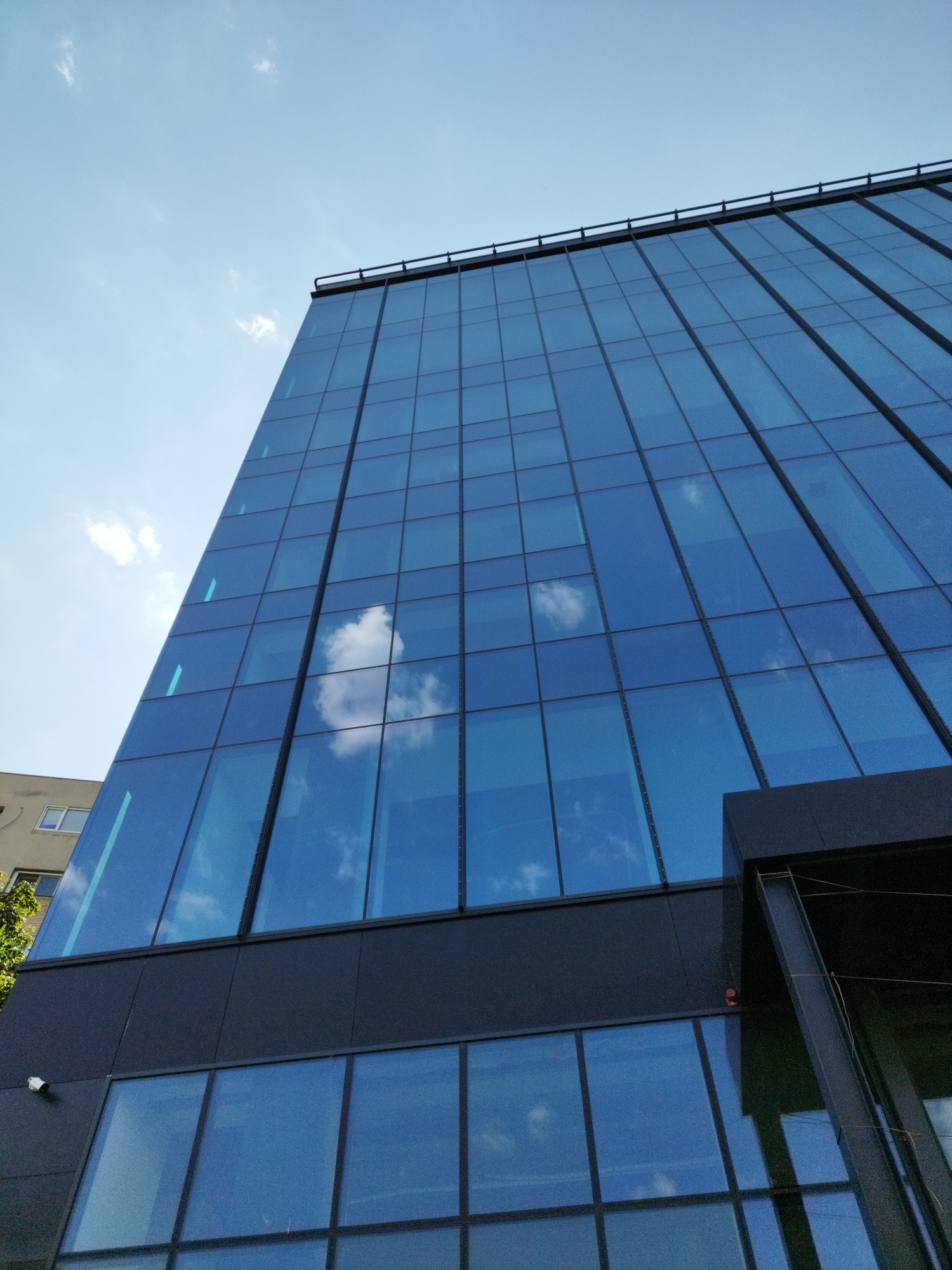The image shows a modern glass-fronted office building against a bright blue sky with scattered white clouds. The building's facade is composed of large, rectangular blue glass panels, reflecting the sky and clouds. A dark, angled section of the building juts out on the right side. To the left, a portion of a lighter-colored, older building with windows is visible, along with some green foliage. A security camera is mounted on the dark facade of the modern building. The overall scene suggests an urban environment, likely during daylight hours. The image is a photograph taken from a low angle looking upwards at the building. There are no people visible in the image.