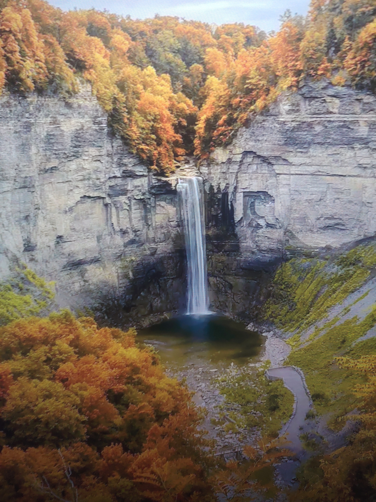 The image shows a waterfall cascading down a high cliff into a pool of water at the bottom. The surrounding environment includes autumnal foliage, with trees exhibiting orange, yellow, and green colors. The cliff walls appear to be made of layered rock formations. At the base of the waterfall, a small stream flows away from the pool. A walking path can be observed on the right side of the image. The location appears to be a natural landscape, possibly a state park, and is located in the city of Unknown, Unknown.