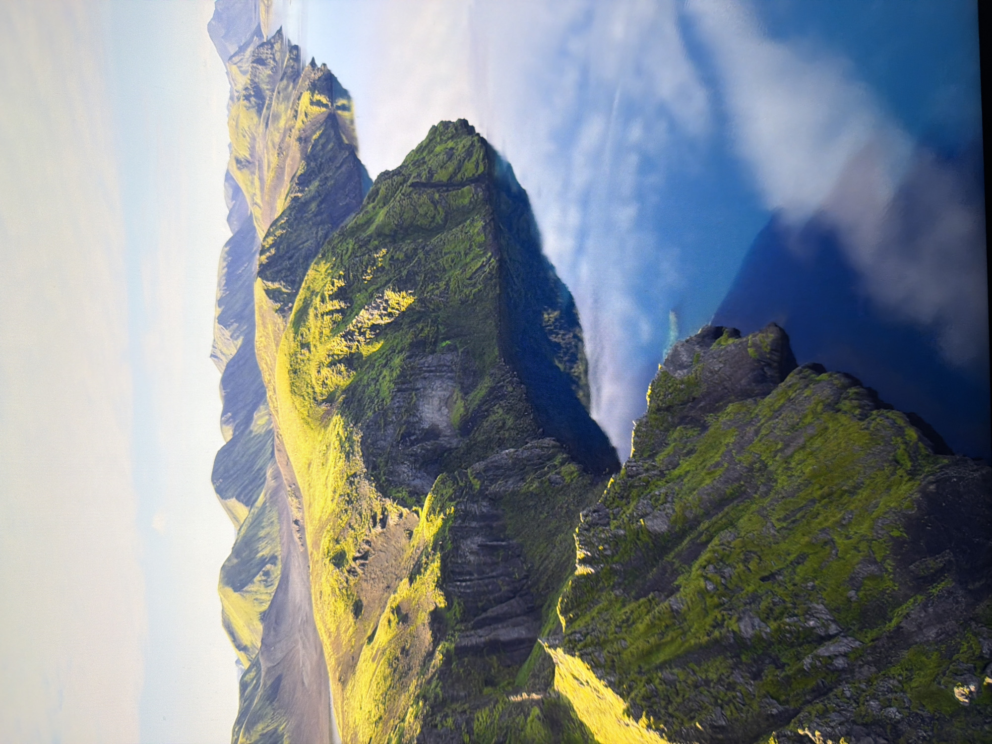 This is an aerial, high-angle, long shot of a dramatic, rugged coastline. The scene is dominated by steep, green, moss-covered cliffs that plunge into a deep blue body of water. The water's surface is calm and reflects the bright sky and scattered clouds. The sunlight highlights the textures of the cliffs, casting long shadows and accentuating the contours of the land. In the distance, a range of rolling, green hills stretches across the horizon under a pale blue sky with wispy clouds. There are no people or man-made structures visible in the image. The location is not in Kyiv, Ukraine; it is a natural landscape.