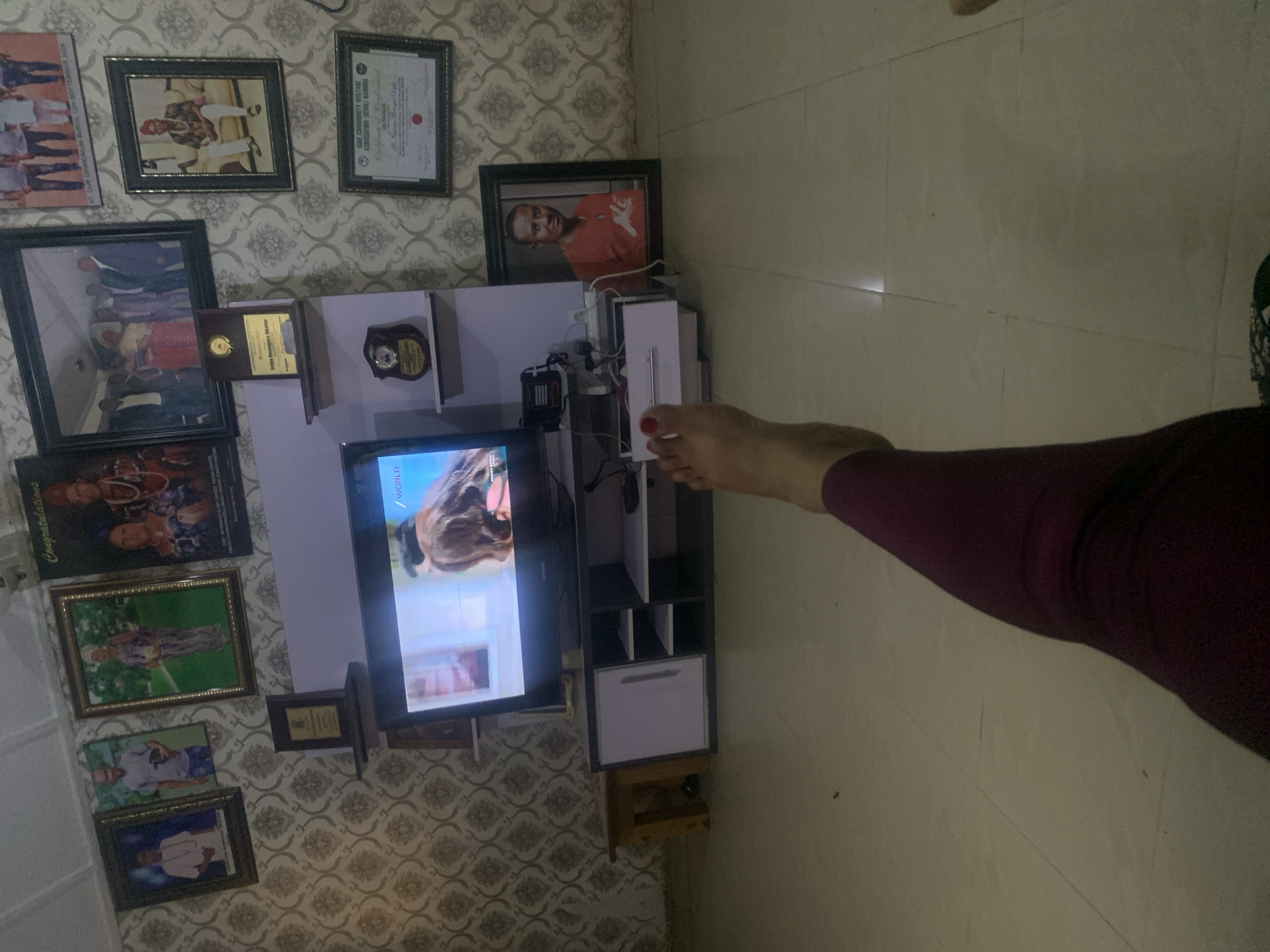 The image depicts an indoor scene, likely a living room, with a focus on framed photographs and a television. The walls are adorned with patterned wallpaper. Several framed pictures are displayed, including portraits of individuals and group photos. One framed certificate is also visible. A television is centrally positioned, displaying an image that appears to be from a movie or show. A modern media console with shelving and cabinets supports the television. In the foreground, a person's left leg and foot are visible, resting in the frame, with painted toenails. The flooring is tiled. The overall impression is of a domestic setting decorated with personal mementos and entertainment. The specific location in Sabon Tasha, Nigeria, is inferred from the context provided.