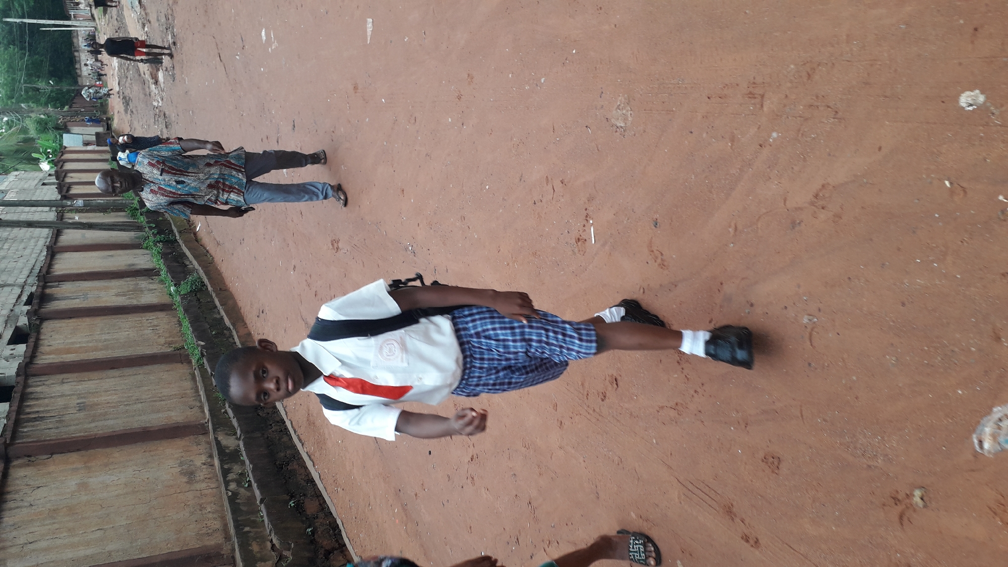 The image depicts a young boy in a school uniform standing on a red dirt road in what appears to be a residential area. The boy wears a white short-sleeved shirt with a red stripe, blue plaid shorts, black shoes, and white socks. He has a backpack slung over his shoulder. In the background, an older man wearing a patterned shirt and jeans is walking away. Other people are visible further in the background. The environment includes a concrete wall with greenery along the side of the road. The scene suggests a street in Onitsha, Nigeria.