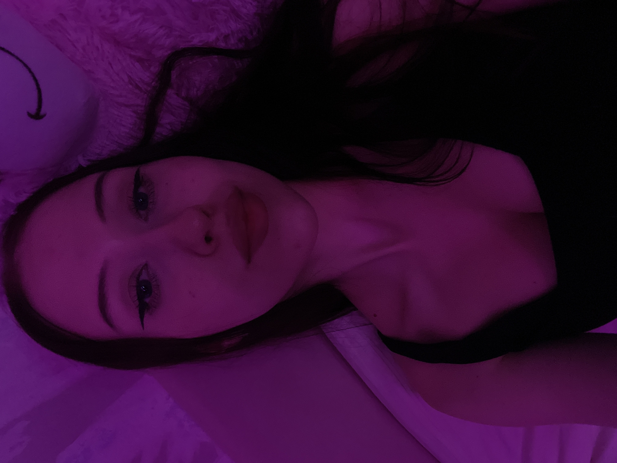 A young woman with dark hair is lying down, possibly in a bed, with a soft, pink, furry object behind her head. The scene is illuminated by purple lighting, casting a magenta hue over everything. The woman is looking directly at the camera, wearing a black tank top. Her makeup includes winged eyeliner and lip gloss. The background appears to be a wall or fabric with a textured surface. There is no indication of the location being Votkinsk, Russia, or any other specific geographic area.