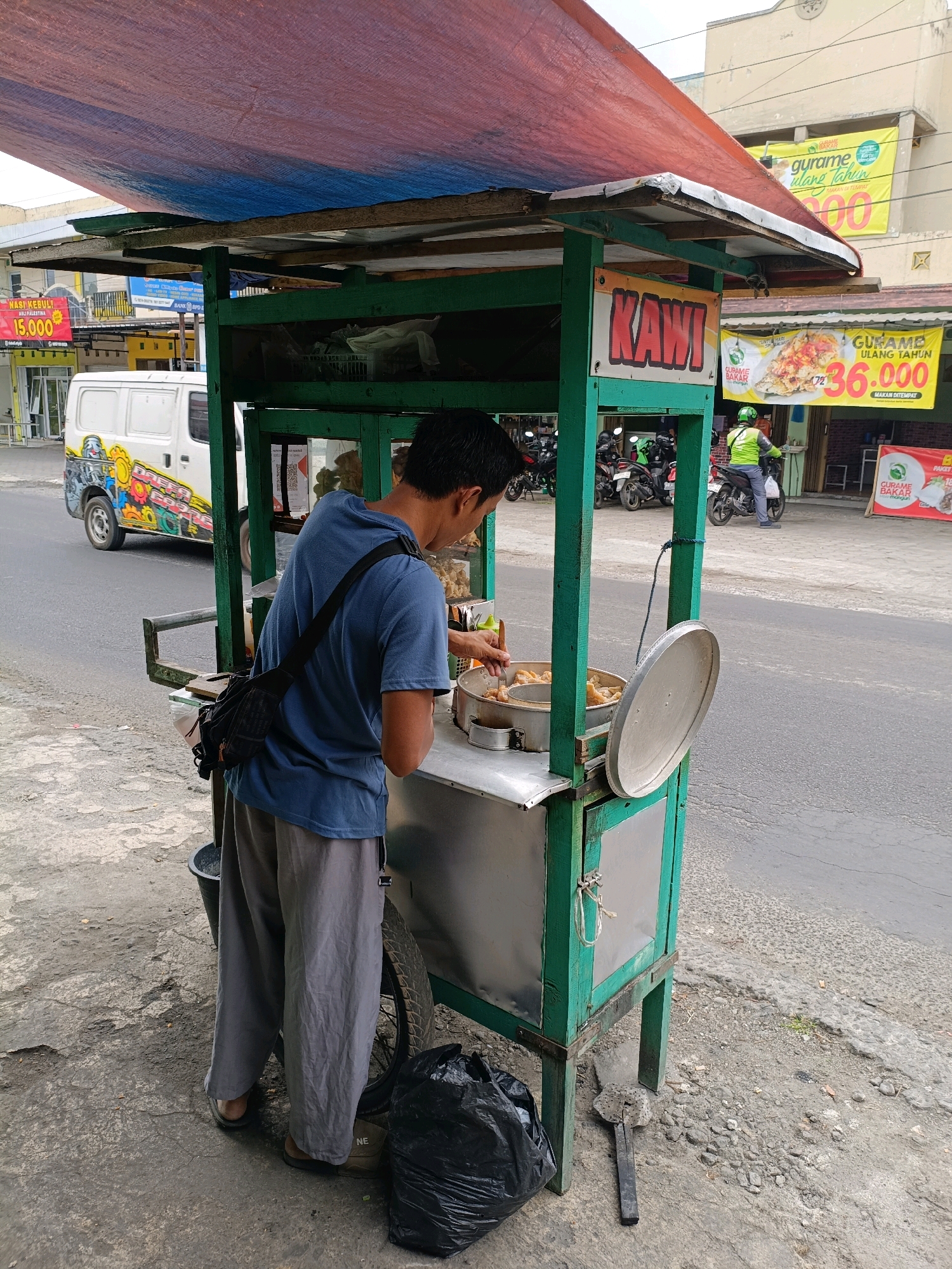 The image shows a street food vendor in Purwomartani, Indonesia. A man, the vendor, is standing behind a green food cart, preparing food. The cart has a red and blue tarp for a roof. In the background, a white van with colorful graphics is parked on the side of the road, and several motorcycles are parked on the sidewalk. There are also banners advertising food prices in Indonesian. The scene is set on a paved street with a sidewalk.