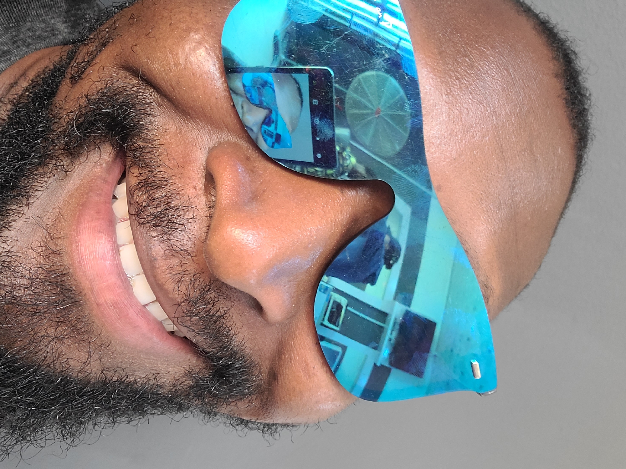 This close-up, sideways portrait captures a smiling man with dark skin and a full, dark beard, featuring some visible grey hairs. The man is wearing distinctive, highly reflective, blue-tinted sunglasses that dominate the right side of his face. His mouth is open in a wide smile, revealing white teeth, which appear to have orthodontic braces or a retainer on the lower teeth.
The sunglasses' mirrored surface clearly reflects elements of the surrounding indoor environment. Prominently visible in the reflection is a mobile phone, held horizontally, capturing its own reflection or a scene beyond. UI elements like a power button and screen icons are discernible on the reflected phone, although no specific text is clearly legible. Also reflected in the glasses is a circular object that resembles a ceiling fan or light fixture, along with geometric patterns and possibly parts of a window or other room features. The background behind the man is a plain, light grey wall.
The image conveys a sense of cheerfulness through the subject's smile. The setting is an indoor space, likely in Jos, Nigeria. The lighting suggests a well-lit environment, but the exact time of day or weather is not determinable. The composition emphasizes the man's facial features and the intricate reflections in his eyewear.