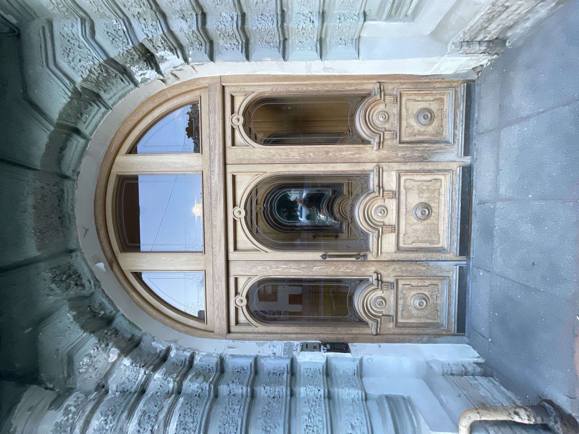 A light brown, double wooden door is set within a stone archway. The door has six glass panels, three on each side, with decorative trim. The top of the door features a large arched window divided into three panes. The stone facade of the building is ornate, with carved details and a textured finish. The area in front of the door is paved with gray flagstones. A metal intercom system is mounted on the left side of the facade, next to the door. The environment suggests a historical building in Saint Petersburg, Russia.