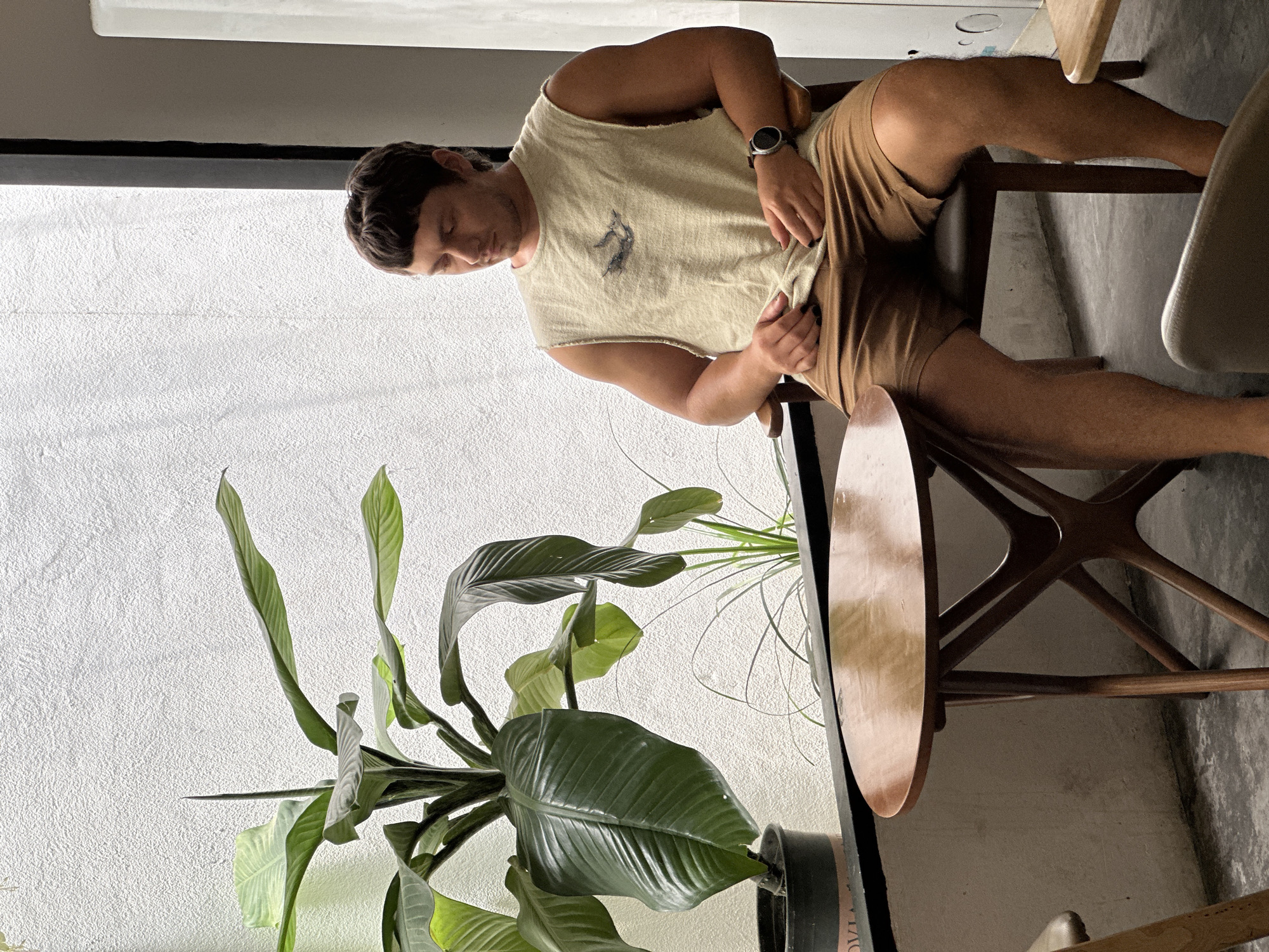 A young man with short brown hair, wearing a light-colored sleeveless t-shirt and brown shorts, sits in a chair at a wooden table. A large potted plant with broad green leaves is to his left. The background is a textured white wall and a window with black trim. This scene is likely in Đà Nẵng, Vietnam, given the indoor plant choice and the casual cafe-like setting.
