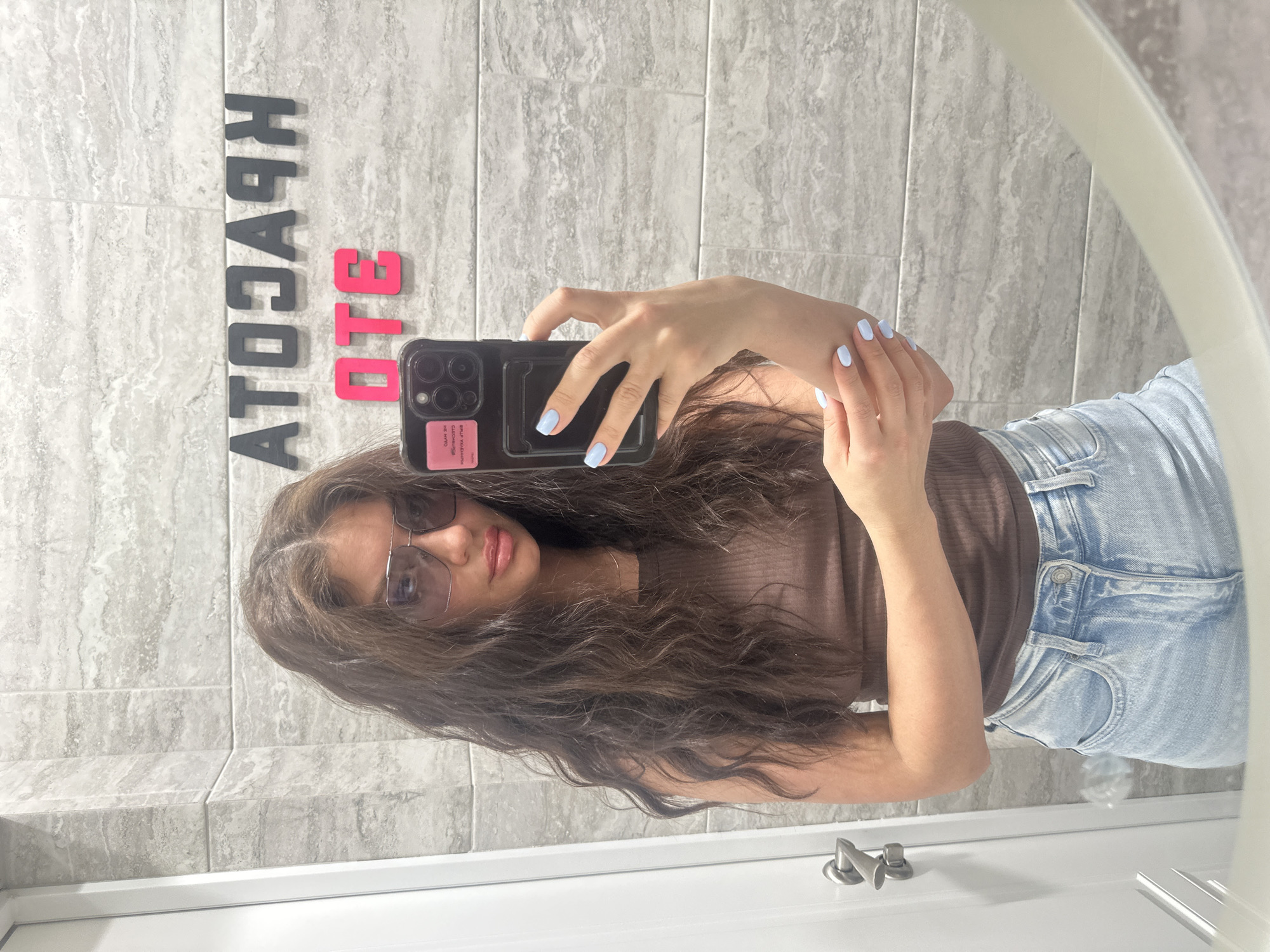 A young woman is reflected in a bathroom mirror. She is taking a selfie with a smartphone. She has long, wavy brown hair and is wearing sunglasses, a brown top, and light-wash jeans. She has light blue nail polish. The bathroom has gray tile walls. On the wall behind her are the backwards Russian words "КРАСОТА ОТЕЦ", possibly "Beauty is the father" . The image does not provide sufficient information to determine the location of the bathroom in Moscow.