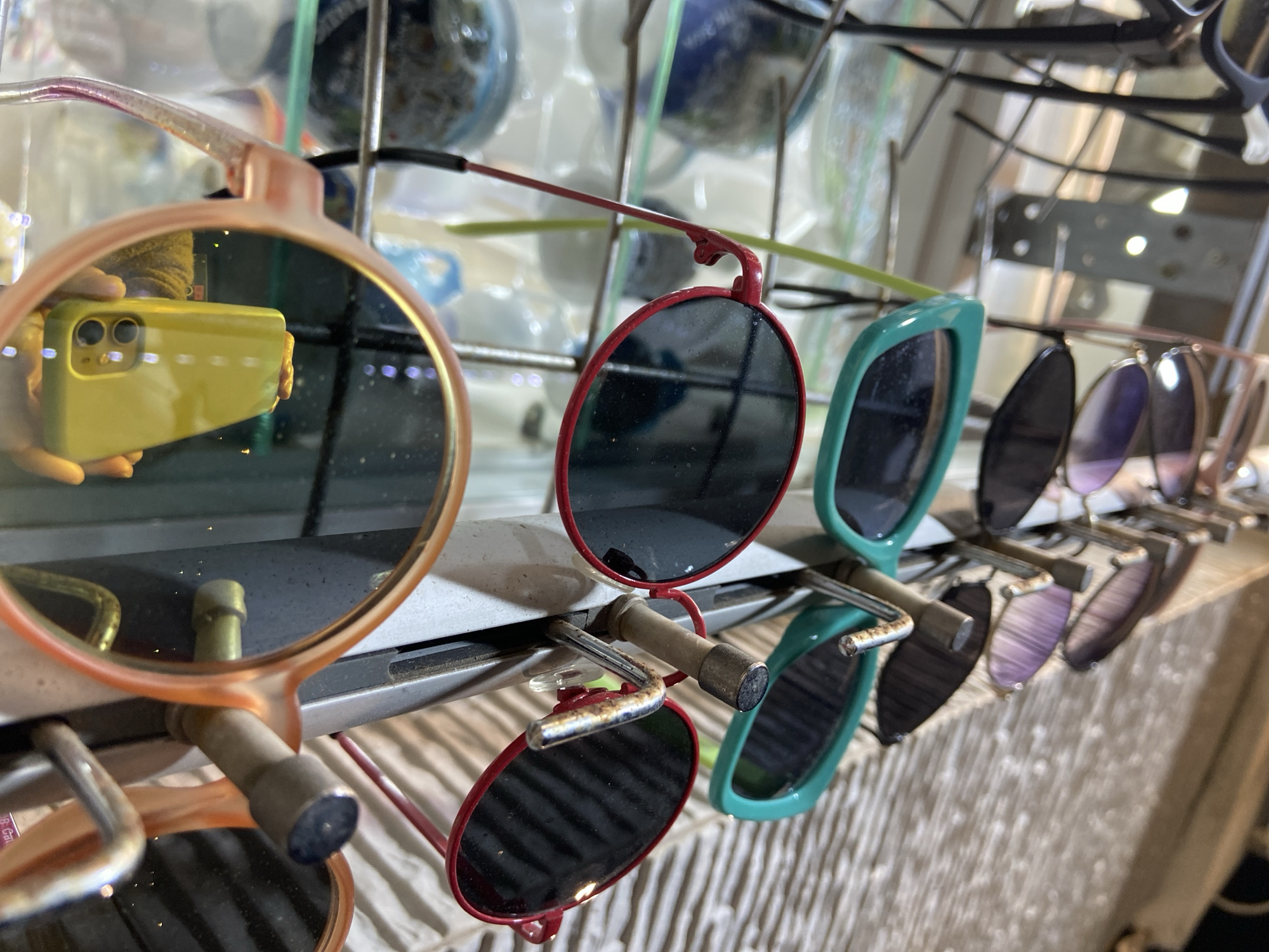 The image displays a close-up, slightly high-angle view of multiple pairs of sunglasses arranged on a metal retail display rack within a retail establishment in Budva, Montenegro. In the foreground, a pair of sunglasses with translucent peach-orange frames and dark, reflective lenses is prominent. The left lens of these glasses reflects a person's hand holding a yellow-green smartphone, indicating the device used to capture the photograph.
Mid-ground subjects include a pair of circular red-framed sunglasses with dark lenses, hanging on a metal peg of the display stand. To its immediate right, another pair of sunglasses features teal-green rectangular frames and dark lenses. The metal display stand exhibits signs of wear, including rust on some of its hooks.
The background shows additional pairs of sunglasses, receding into blur, with visible frames and lenses ranging from dark to purple-gradient. Blurred elements in the periphery suggest other merchandise or ambient light sources, and a ribbed or textured surface is partially visible in the upper right. No other individuals are present within the frame beyond the reflection.
