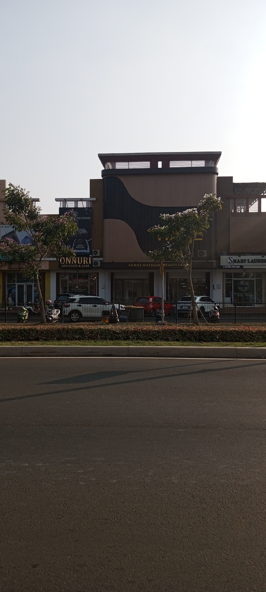 The image shows a street view of a commercial building in Lambangjaya, Indonesia. The building features a modern architectural design with a prominent dark, wavy panel. Several storefronts are visible, including "ONNURI BROWNIES & CAKE" and "SMART LAUNDRY". Cars and motorcycles are parked or passing by on the street in front of the buildings. Trees and bushes line the sidewalk. The sky is clear and bright, suggesting daylight.