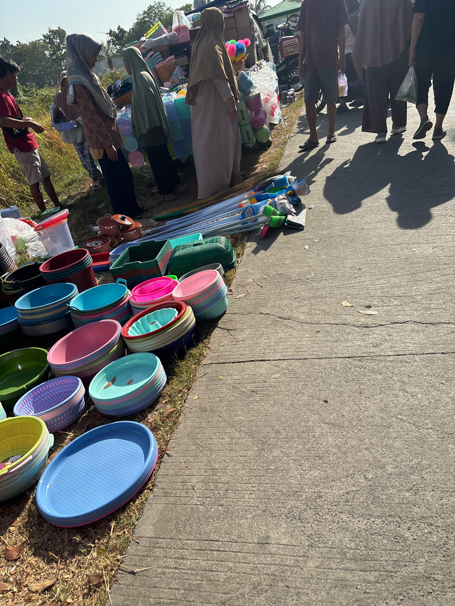 This image depicts an outdoor market scene in Telagamurni, Indonesia, with a strong focus on the sale of household items, primarily plastic basins and kitchenware. A group of people, some wearing hijabs, are visible, engaged in either shopping or selling. Several women in hijabs are positioned around stalls displaying a variety of colorful plastic basins stacked in neat rows. One woman in a light brown hijab stands with her back to the camera, while another in a grey hijab is turned sideways, appearing to interact with someone off-frame. A young man in a red shirt and shorts is also present, standing on the left side of the image. The ground is a mix of dry grass and a concrete pathway, on which more items, including brooms, are laid out. In the background, there are glimpses of structures and possibly vehicles, suggesting a bustling marketplace environment. The lighting indicates it is daytime, with shadows cast on the concrete, suggesting a sunny day. The overall impression is a typical local market scene, offering everyday goods to the community.