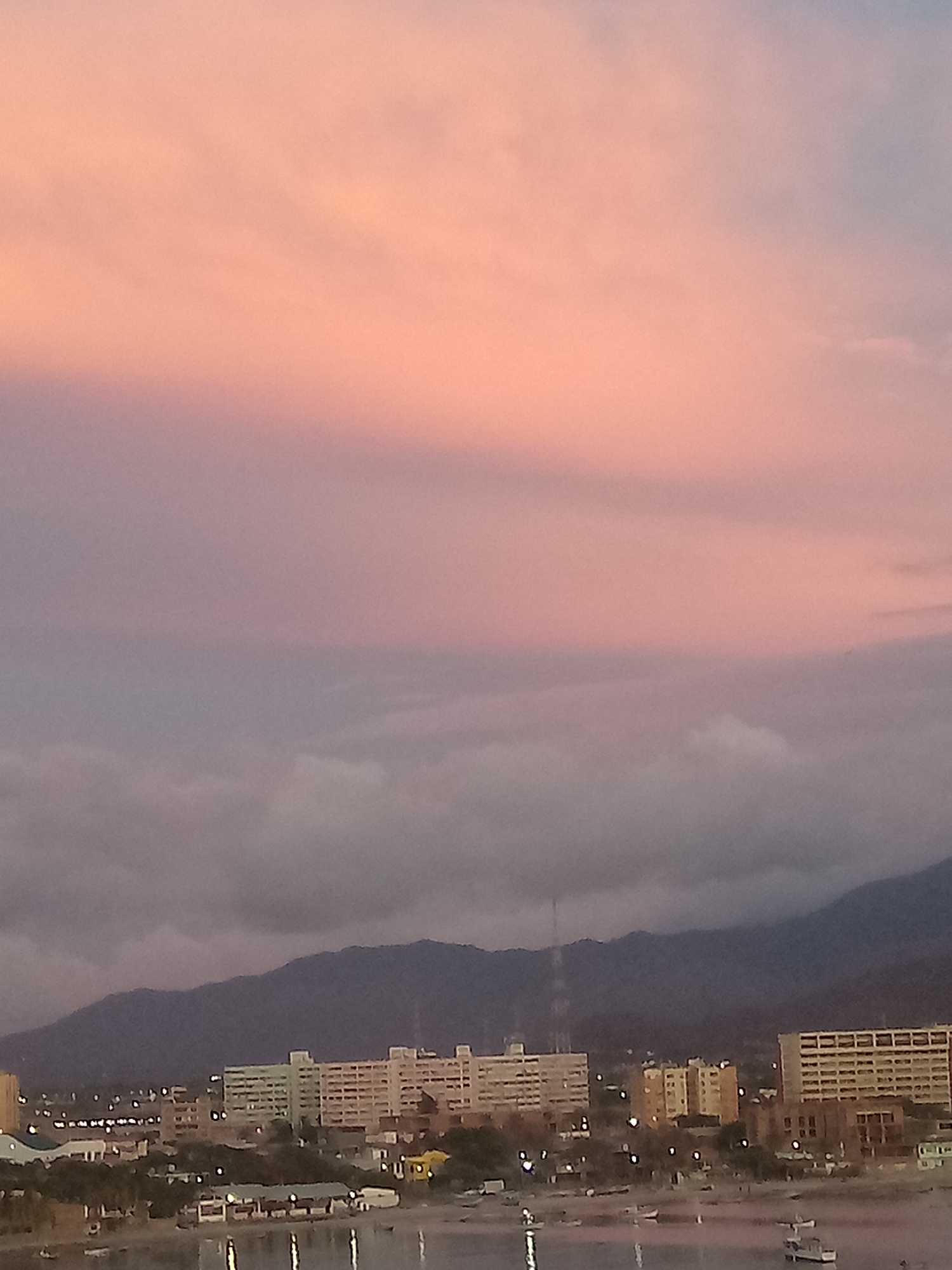 The image shows a cityscape at dusk in Juan Griego, Venezuela. In the foreground, a body of water reflects the lights of the city. Behind the water, a cluster of buildings stands, illuminated by streetlights. A mountain range forms the backdrop to the city. The sky is a gradient of color, transitioning from a pinkish hue at the top to gray clouds near the horizon. A tall, slender structure, possibly a telecommunications tower, rises from behind the buildings. Small boats dot the water.