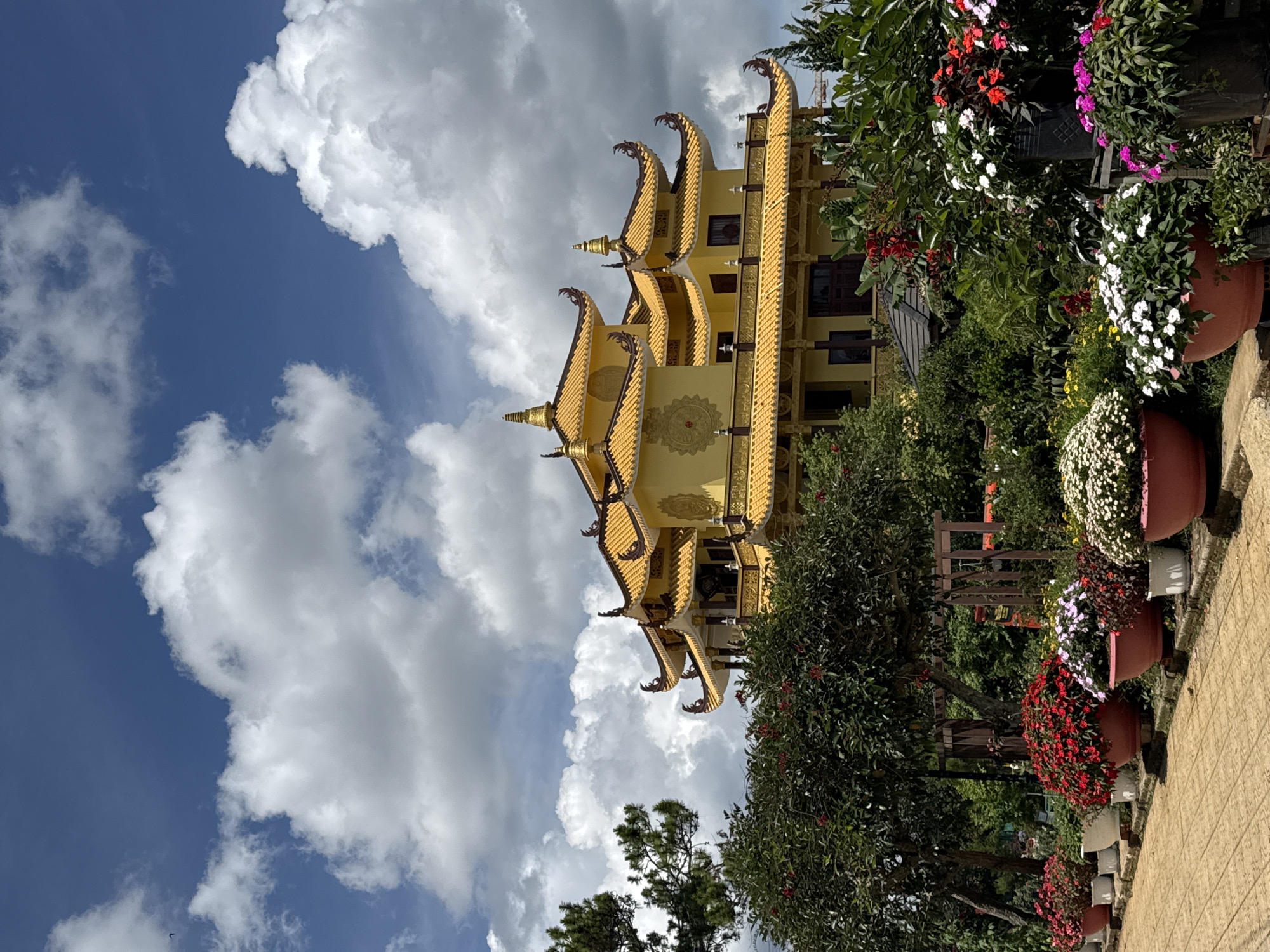 The image shows a yellow, multi-tiered building with ornate rooflines against a backdrop of blue sky and fluffy white clouds. The building is located in Da Lat, Vietnam. Lush greenery and potted flowers frame the building on its right side. A paved path is visible at the bottom right corner. There are no people visible in the image.