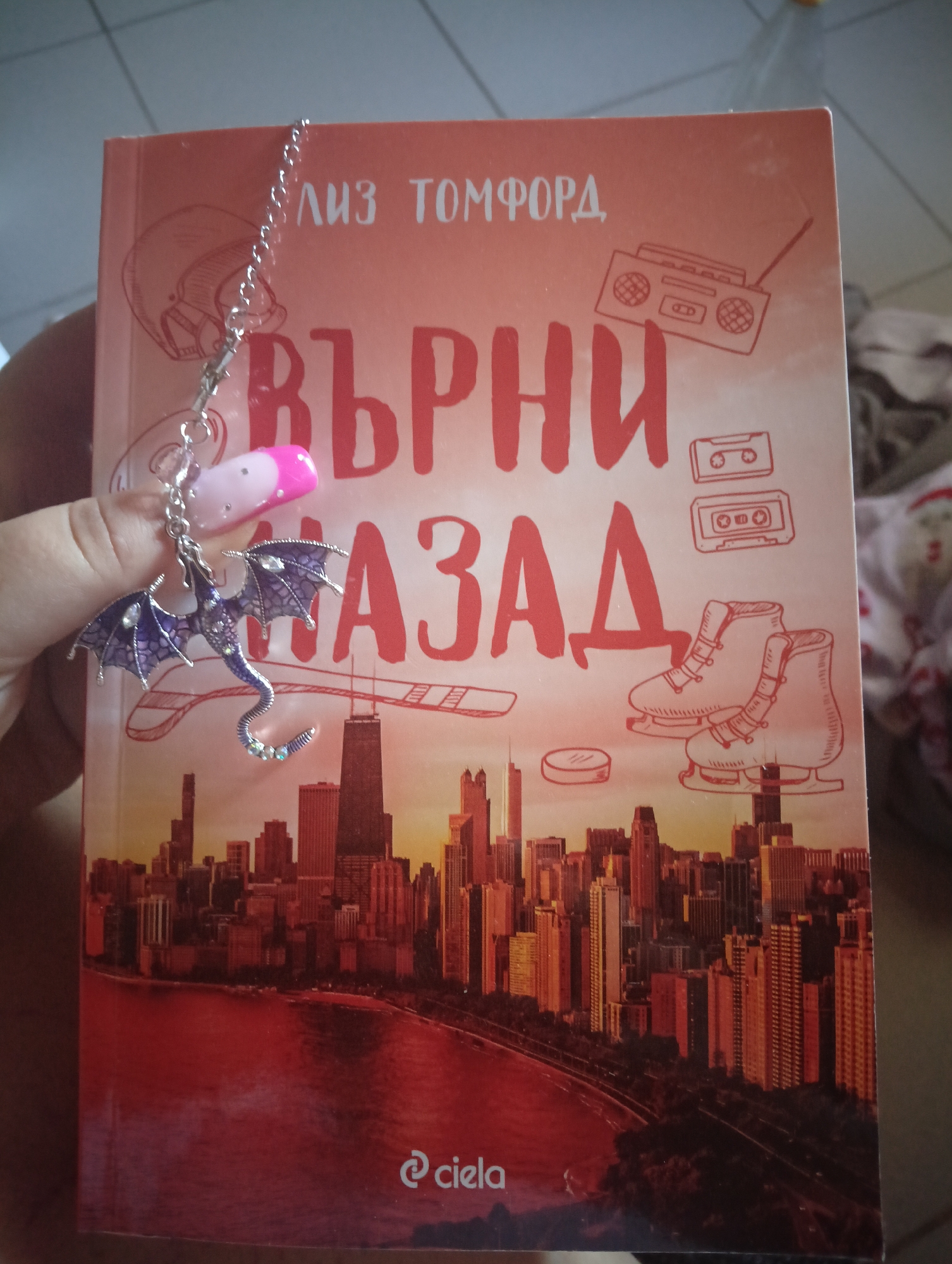 A book with a pink cover is being held up by a hand with pink and white nails. The book title is "Върни Назад" by Лиз Томфорд. The cover features a cityscape, possibly Chicago, with the lake in the foreground. Above the city, there are drawings of ice skates, cassettes, and a boombox. A dragon-shaped bookmark is attached to the top of the book. The bottom of the book has a logo that says "ciela".