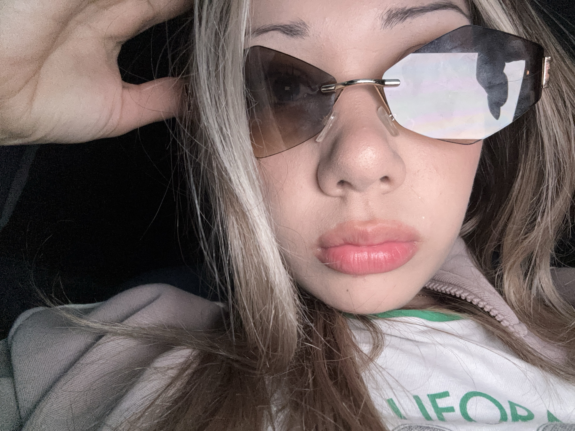 A close-up, eye-level selfie captures a young adult, likely female, in Dobrich, Bulgaria. The individual has light skin, full pink lips with a glossy finish, and dark, well-defined eyebrows. Her hair is long, featuring prominent blonde highlights mixed with darker brown strands, with some hair falling over her shoulders and chest. She is wearing frameless, hexagonal or octagonal sunglasses with gold-toned bridge and temple pieces. The left lens (viewer's right) is dark brown with a gradient, while the right lens (viewer's left) is highly reflective, showing a distorted dark shape. Her left hand, with fingers slightly bent, is raised and lightly touches the side of her head near her temple. She wears a light-colored, possibly off-white or light grey, zippered top, likely a hoodie or sweatshirt, with a visible light pink zipper and a green collar or piping underneath. Partially visible green text on the fabric reads "LIFO" or "LIFOR". The background is dark and out of focus, providing no specific environmental details.