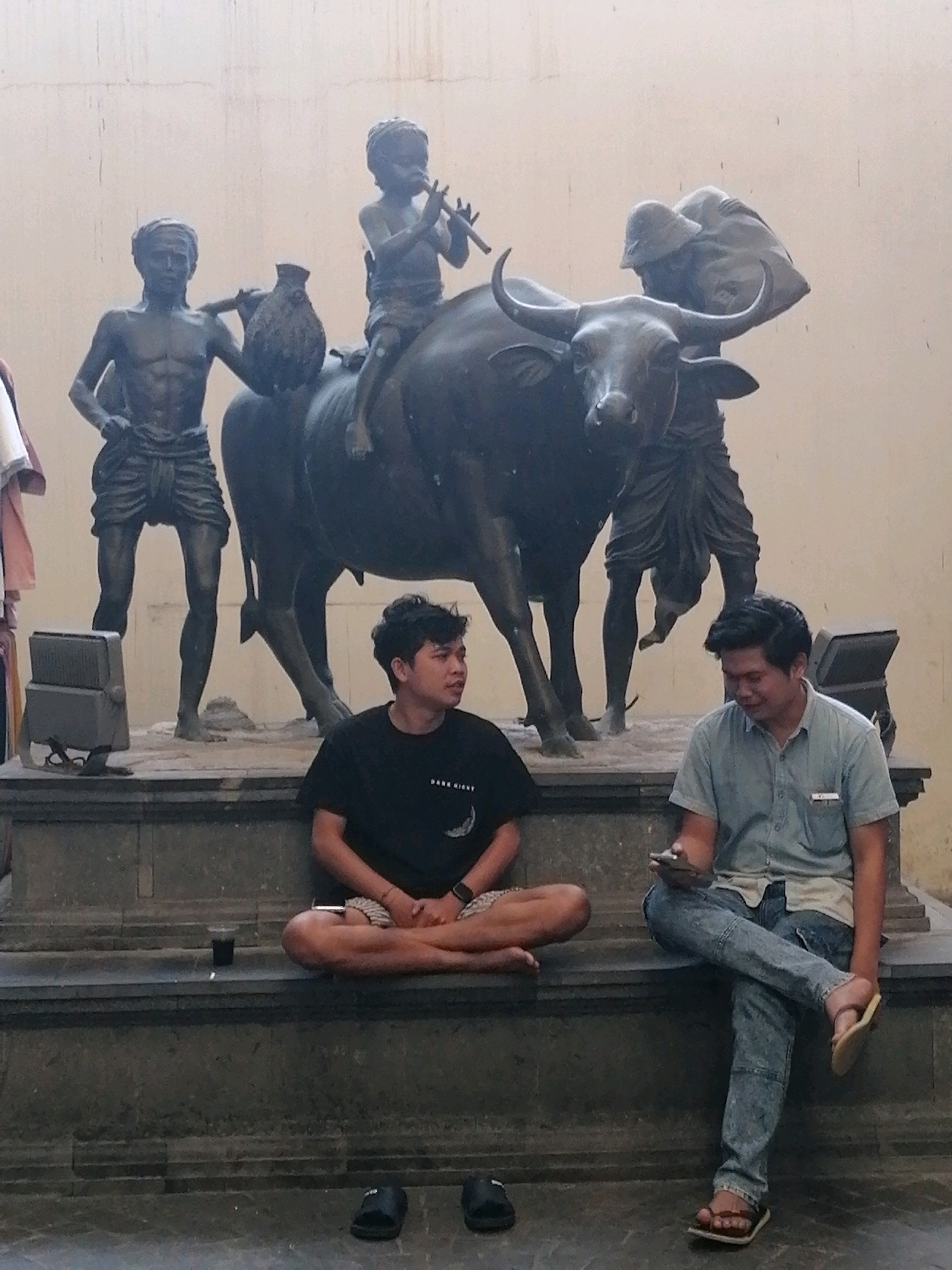 In Ubud District, Indonesia, two men are seated on a stone platform in front of a bronze sculpture. The sculpture depicts a water buffalo with three figures. One figure, a child, sits on the buffalo's back, playing a flute. Another figure stands to the left, holding a large pot. A third figure stands behind the buffalo, with their head covered.
The man on the left is seated cross-legged, wearing a black t-shirt and patterned shorts. He has a cup of dark liquid on the platform next to him. The man on the right is wearing a light blue short-sleeved button-down shirt and distressed blue jeans. He is looking down at a smartphone in his hand and is wearing flip-flops. The background is a textured wall. To the left of the sculpture, there are some hanging garments. Two floodlights are mounted on the platform. A pair of black flip-flops are on the ground in the foreground, with another pair to the right.