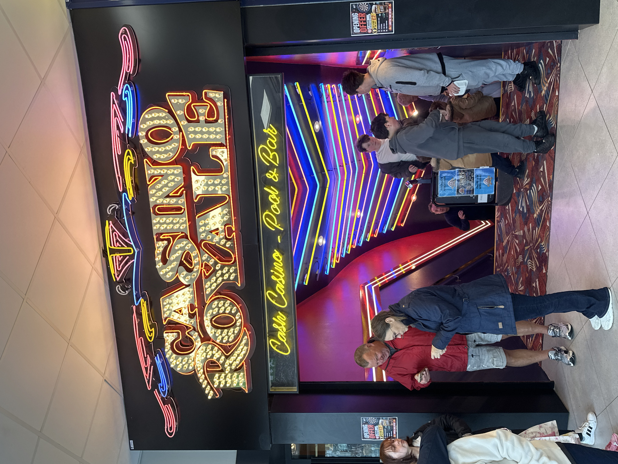 This image depicts the brightly lit entrance to "Casino Royale Amusements" located on Marine Parade in Great Yarmouth, United Kingdom. Above the entrance, a large, retro-style neon and bulb sign displays "CASINO ROYALE" in prominent yellow and red letters, topped with additional decorative neon elements in pink, yellow, and blue. Below this, a yellow neon sign reads "Cash Casino - Pool & Bar." The interior of the entrance features vibrant, multi-colored horizontal and diagonal neon light strips in blue, pink, and yellow, illuminating purple and black wall sections. The floor inside is covered with a patterned carpet, while the exterior foreground is tiled.
Approximately ten individuals are present around the entrance. In the center-left, a man in a red jacket and light shorts stands, looking at a mobile phone in his hand. Adjacent to him, a woman in a dark blue jacket and dark trousers, wearing glasses, also looks down at an object in her hands. To their left, a young woman with brown hair and a dark jacket looks towards the entrance. Partially visible in the far left foreground is a person in a light top and dark trousers wearing white Adidas sneakers.
Inside the entrance, on the right, a young man in a grey hoodie and sweatpants holds a white object while another young man in similar attire stands looking down. Further inside, a third young man in a white top and dark jacket is partially visible. In the deeper background, a man in a dark shirt is faintly seen. On either side of the entrance, black boards display "OPENING OFFER" promotions; the right poster includes details about discounted pool table usage and mentions "FAMILY WELCOME" and "FAMILY AMUSEMENTS." A blue-framed stand inside the entrance also displays similar "FAMILY WELCOME" signage.