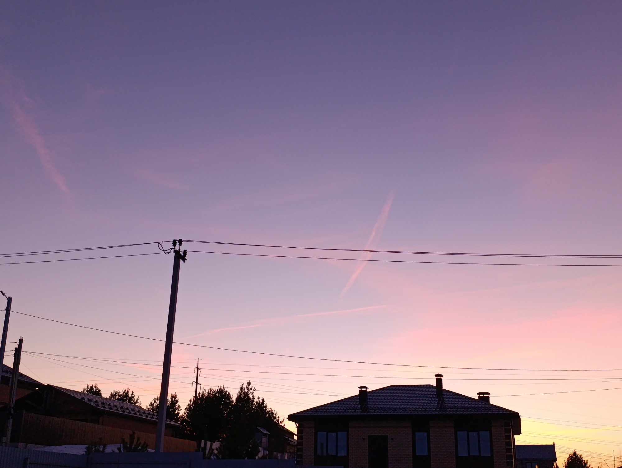 The image shows a residential area in Chaikovsky, Russia at dusk or dawn. The sky transitions from a deep purple at the top of the frame to a soft pink and orange near the horizon. Two contrails intersect in the sky. A house with a dark roof and brick exterior is visible in the lower portion of the image, along with power lines and utility poles. Some dark trees are silhouetted in front of other houses. The overall scene depicts a quiet, suburban setting.