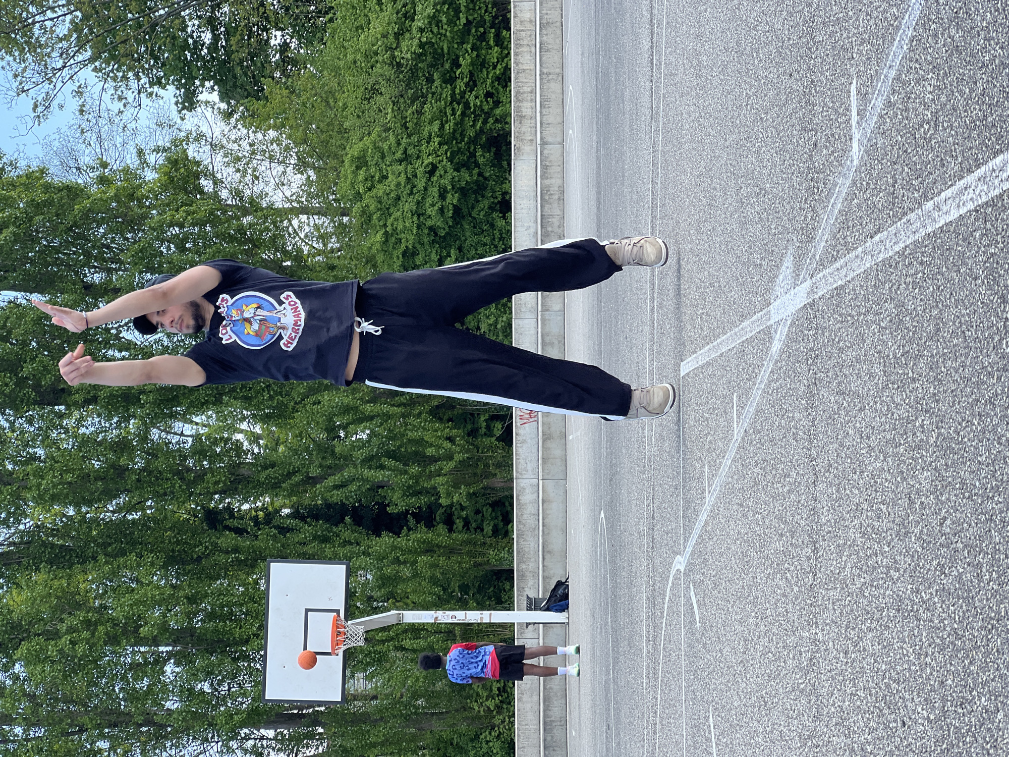 An outdoor, slightly rotated image depicts an open-air basketball court flanked by lush green trees and a concrete wall or steps, under a bright sky. The scene is likely in Strasbourg, France, as per the provided context.
In the foreground, a man with a light complexion, wearing a black t-shirt, black track pants with white stripes, and light-colored sneakers, is striking a pose. He faces the camera, his arms are outstretched to his sides with palms open, and he wears a baseball cap backward. On his t-shirt, a circular logo is visible with cartoon characters and the text "LOS HERMANOS" and "SONNTAG ABEND".
Further back on the court, another person, seen from behind, stands near a basketball hoop. This individual wears a colorful red, blue, and white patterned t-shirt, black shorts, and bright green socks with white sneakers. A basketball is visible inside the net of the hoop, suggesting an ongoing or recently concluded game. A dark backpack or bag rests on the concrete steps behind this person.
The court itself is made of asphalt or similar material, marked with worn white lines. The surrounding trees are full of green foliage, indicating a warm season. The lighting suggests it is daytime, possibly late morning or early afternoon, with diffused light under an overcast or hazy sky.
