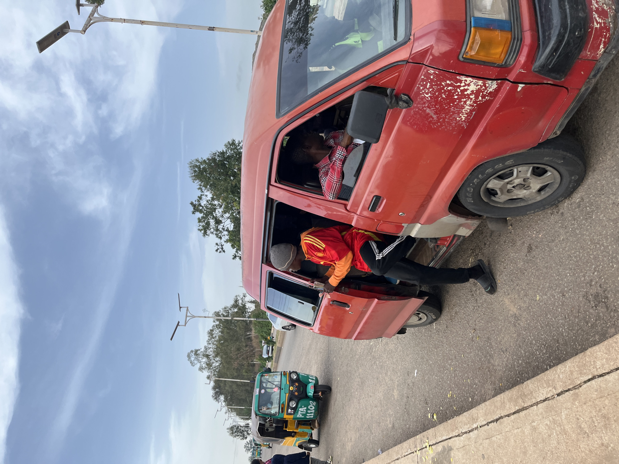 The image depicts a street scene in Gura Top, Nigeria, with a red minivan as the central subject. A young man wearing a gray beanie, a red and yellow jacket, and black pants with white stripes is standing in the open doorway of the minivan, holding onto something inside. He is facing towards the right of the frame, with his left foot on the ground. Another person, wearing a red and black checkered shirt, is visible in the driver's seat of the minivan, looking out the window to the right. The minivan appears weathered, with peeling paint on its exterior.
To the left of the minivan, a green and yellow auto-rickshaw is parked on the road. The rickshaw has "PTA 11102" printed on its side. Another person is seated in the driver's seat of the rickshaw. Further down the road, several trees and streetlights are visible. The sky is partly cloudy, with a light haze. The road appears to be paved, with a concrete curb and some scattered leaves on the ground. The overall atmosphere suggests a typical day in a Nigerian urban or semi-urban environment, likely a transportation hub given the presence of both a minivan and an auto-rickshaw.