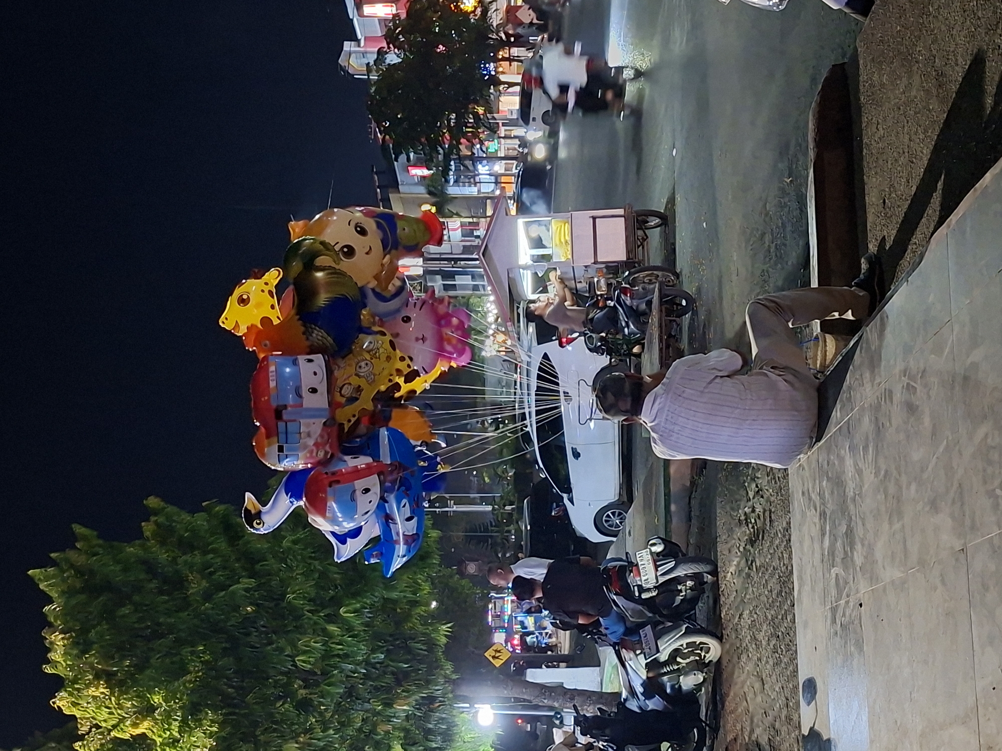 A nighttime scene in Lhokseumawe, Indonesia shows a man sitting on a ledge, possibly a curb, wearing light-colored pants and a pale, short-sleeved button-down shirt. He appears to be observing activity on the street. To the left, a large cluster of character-shaped balloons in various colors (yellow, orange, blue, pink) is tied together. In the background, there's a brightly lit street with people on scooters and other vehicles. A food cart with a vendor is positioned on the side of the road. There are also several motorcycles parked near the curb. Buildings with lights line the street in the background, and a tree is visible on the left. The ground appears to be a sidewalk or paved area.