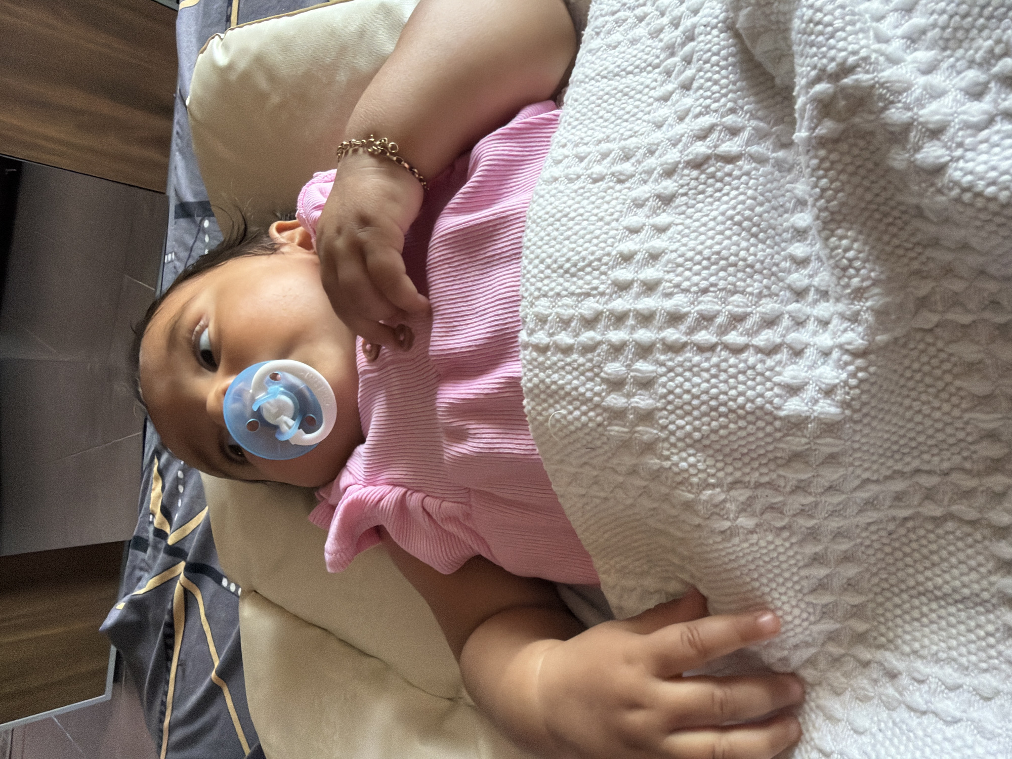 An infant, wearing a pink ribbed shirt and a pacifier, lies on a white textured blanket with a gold and gray patterned sheet underneath. The infant is on a bed, indicated by the pillow and bedding. A golden bracelet adorns the infant's left wrist. The background shows wooden furniture. The image is taken indoors, suggesting a domestic setting. The location is described as Razhevo Konare, Bulgaria.
