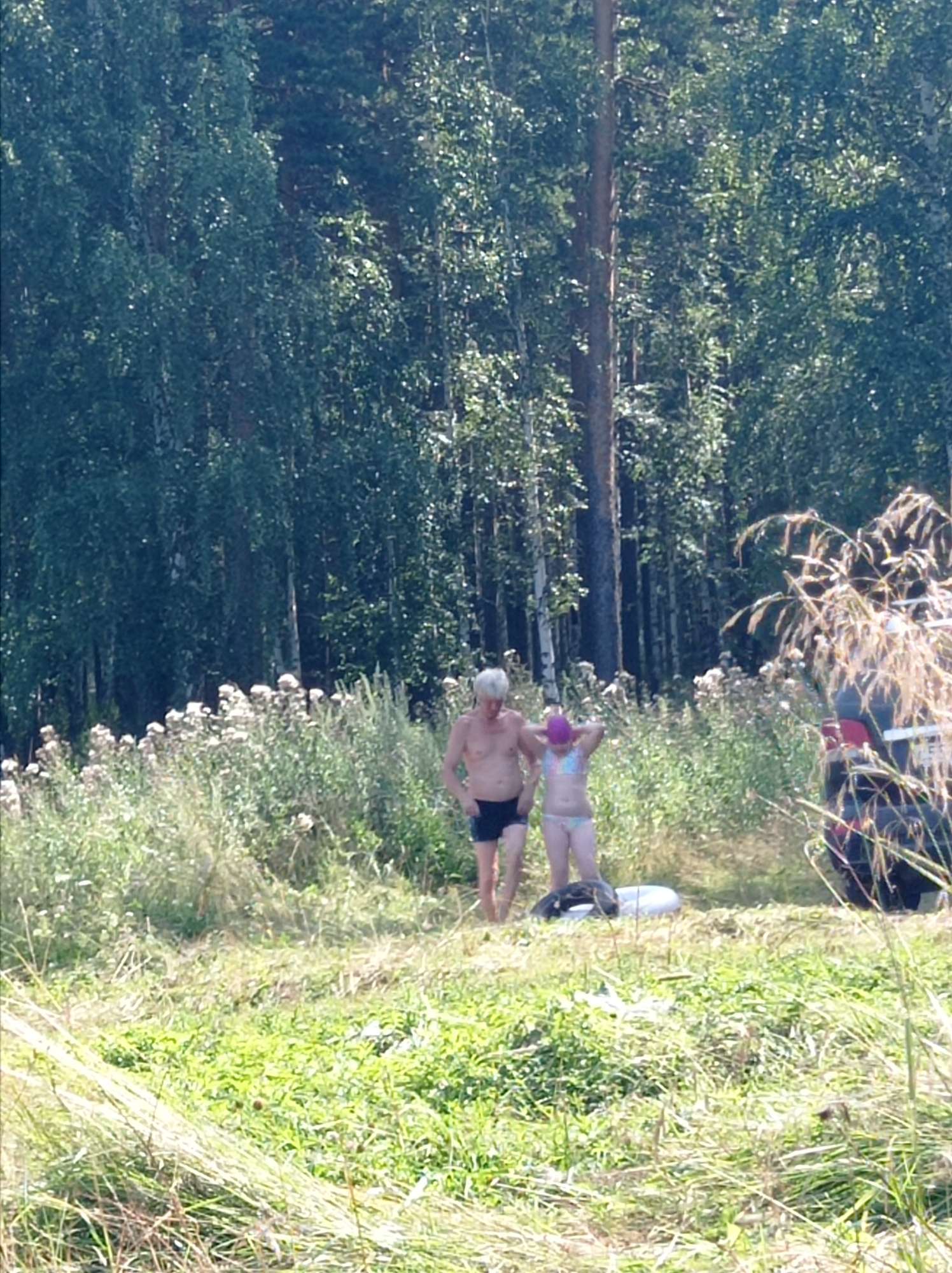 Two people, a man and a woman, are standing in a grassy, overgrown area with a forest in the background. The man, on the left, is wearing dark blue swim trunks. The woman, on the right, is wearing a patterned bikini and a purple swim cap. They appear to be outdoors, possibly near a body of water given the presence of inflatable rings on the ground. A dark-colored vehicle is partially visible on the right side of the image. The environment suggests a rural or semi-rural setting, characteristic of areas surrounding cities like Degtyarsk, Russia, which is known for its natural landscapes.