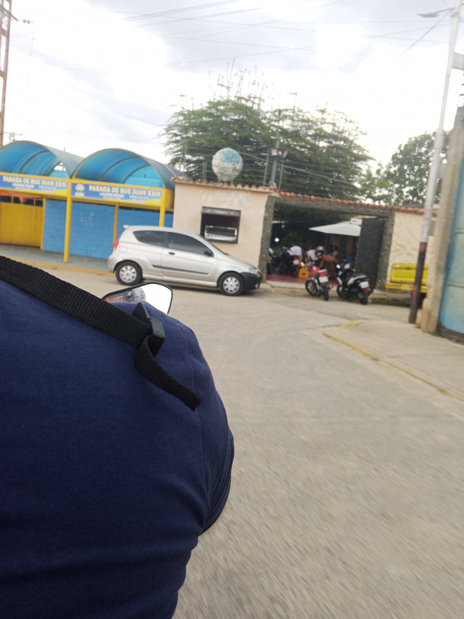 The image, taken from a moving vehicle (likely a motorcycle) in Tinaquillo, Venezuela, features a partial view of the driver's shoulder with a blue shirt and black strap in the foreground. In the midground, a silver hatchback car is parked next to a yellow and blue structure labeled "PARADA DE BUS JUAN XXIII" (Bus Stop John XXIII). Adjacent to this structure is a beige building with a covered area where several people are seated and motorcycles are parked. The background includes trees and a cloudy sky. The street surface and other buildings are also visible, suggesting an urban or suburban setting.