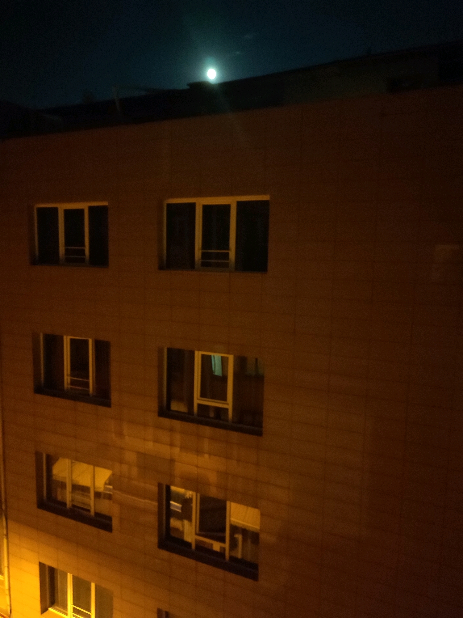 This is a nighttime, eye-level, medium shot of a multi-story building with a tiled facade in Kadıköy, Turkey. The sky above is dark blue, with a bright, full moon visible towards the upper right. The building has multiple windows, some of which are illuminated from within, casting a warm yellow glow. The windows appear to be rectangular and are arranged in rows. The overall impression is one of urban nighttime quietude.